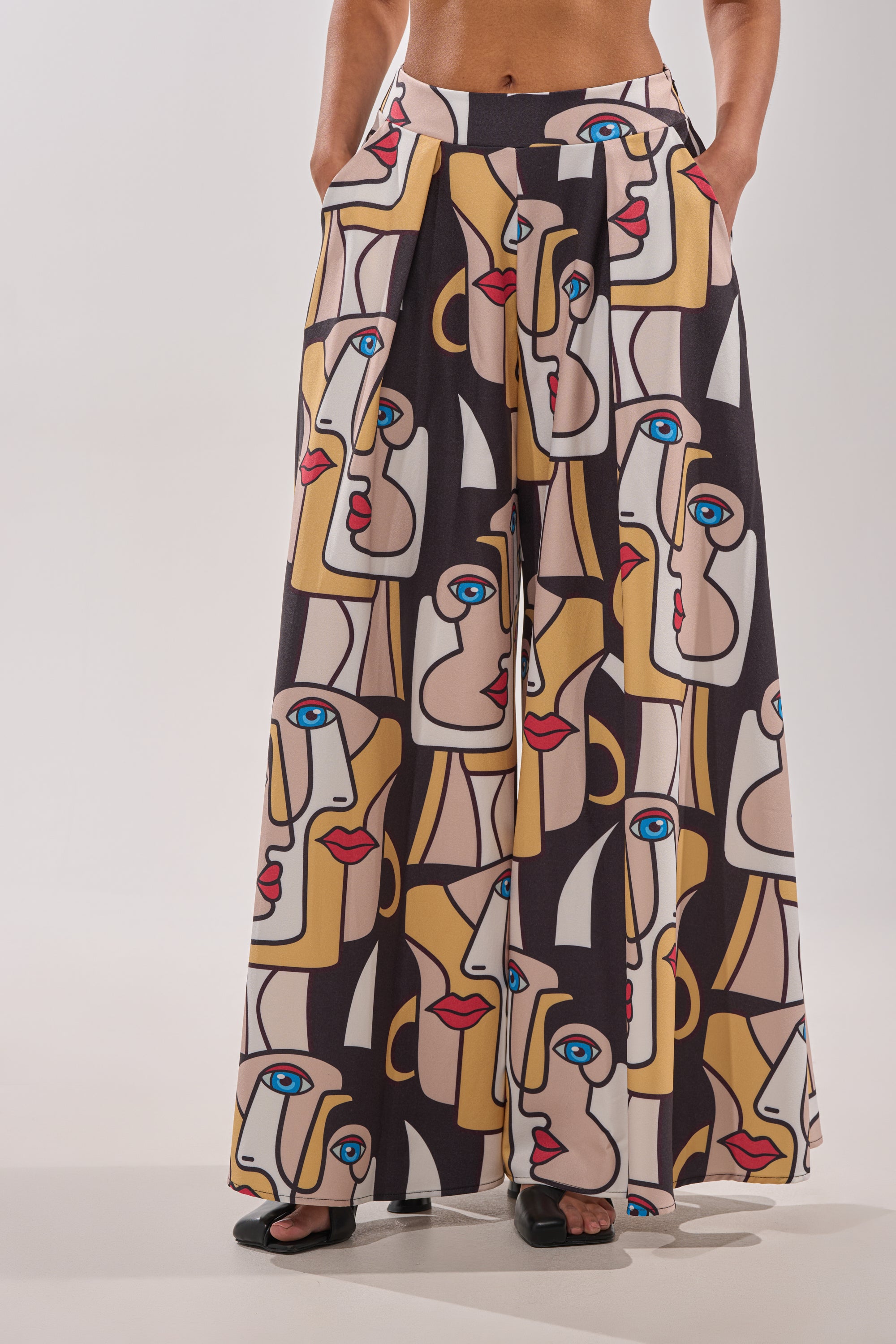 A person stands barefoot with hands in pockets, wearing the FACES EVERYWHERE TROUSER—wide-leg pants with a bold face print in yellow, beige, black, red, and blue—against a plain background. 1