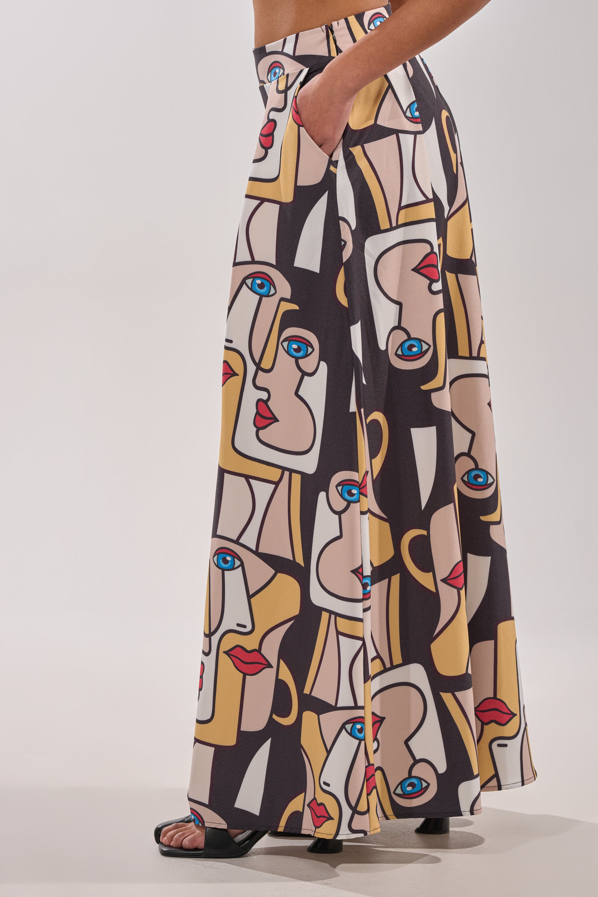 A person wears the FACES EVERYWHERE TROUSER—wide leg, high-waisted pants with a colorful abstract face print—standing sideways with one hand in their pocket and black open-toe heels. 3
