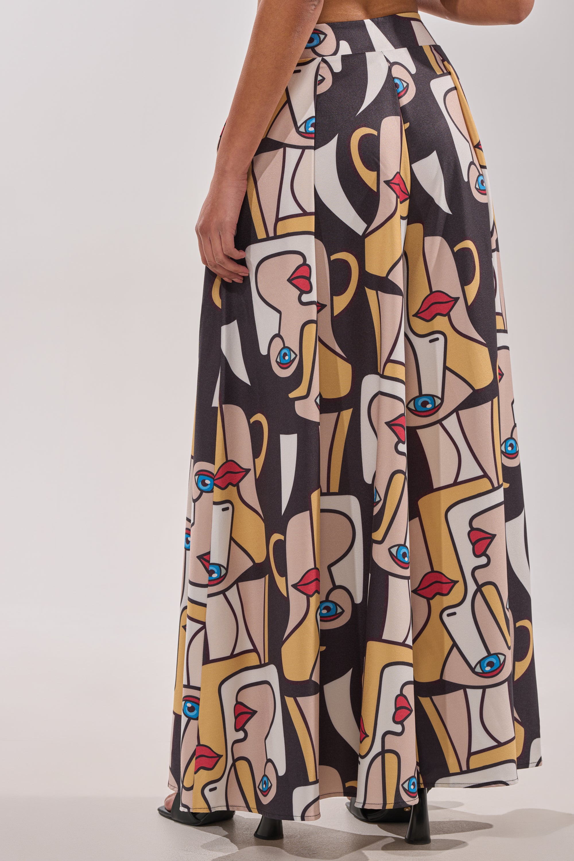 A person stands against a plain light background wearing the FACES EVERYWHERE TROUSER, featuring a bold abstract face pattern in beige, yellow, black, red, and blue tones. 2
