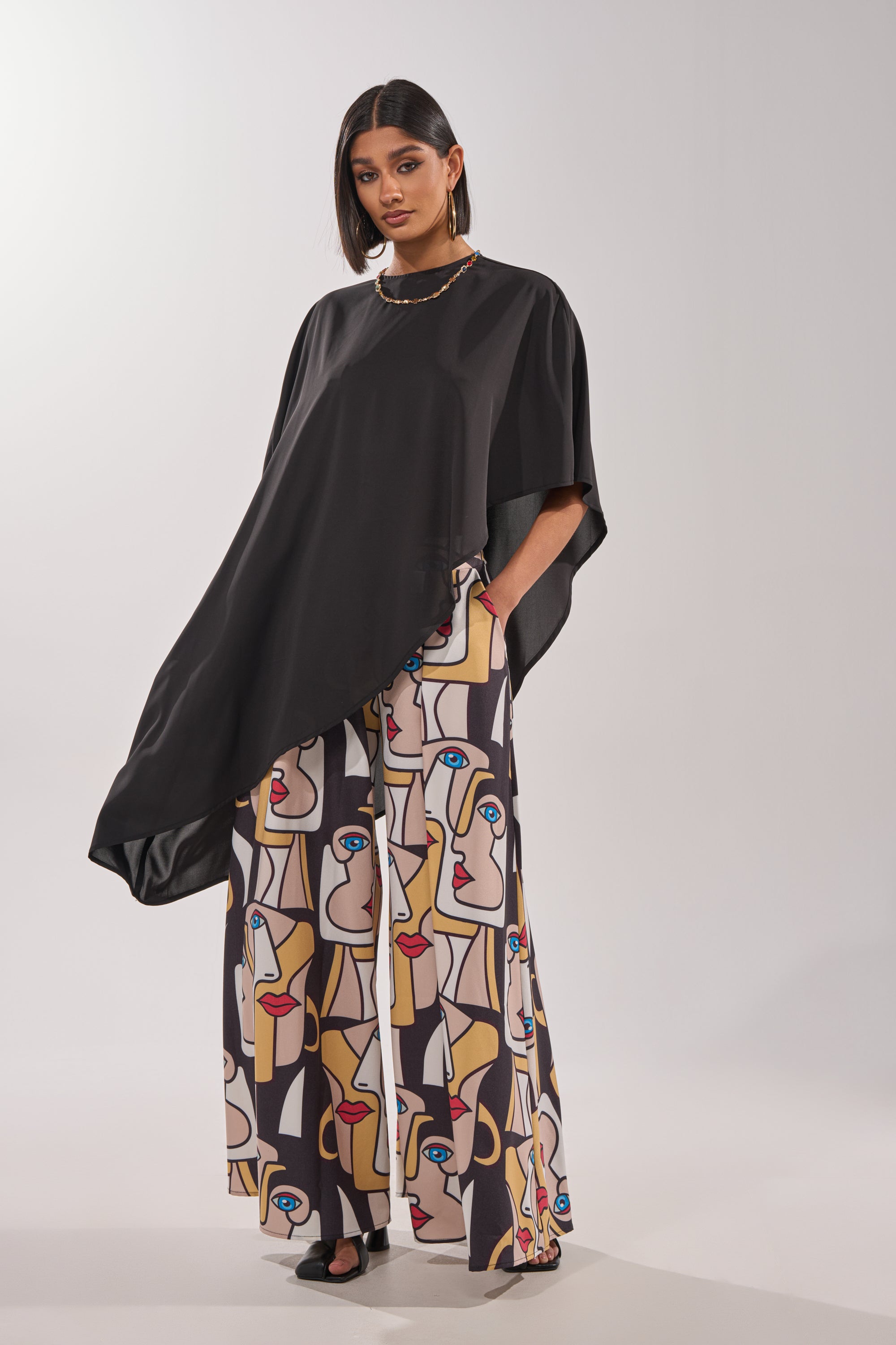 A woman wears the FACES EVERYWHERE TROUSER, featuring a colorful abstract face print, paired with a loose black asymmetrical top. She completes her look with hoop earrings, a necklace, and black sandals against a plain background. 4