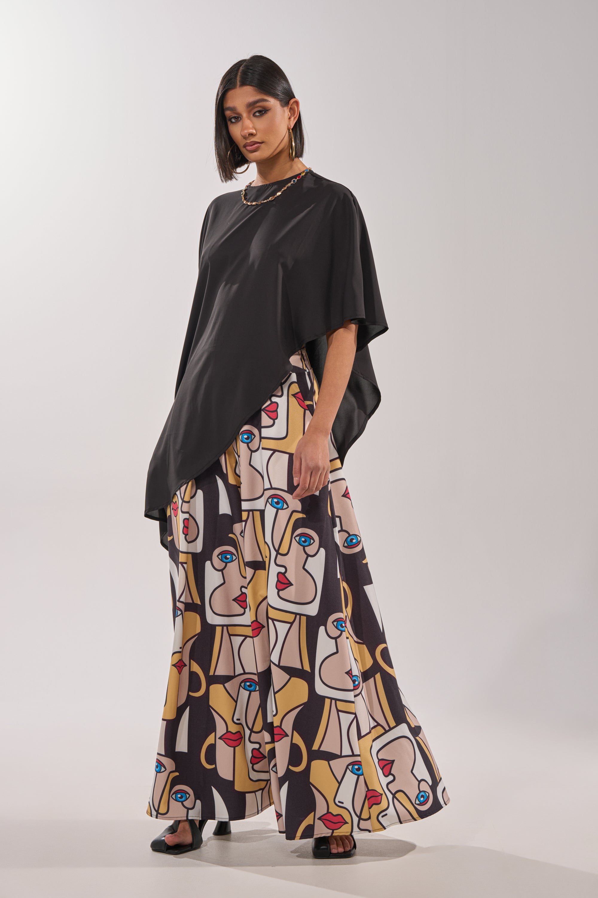 A woman wears straight, shoulder-length hair, black sandals, a loose black top, and the FACES EVERYWHERE TROUSER featuring an abstract face print in beige, yellow, red, blue, and white. 5