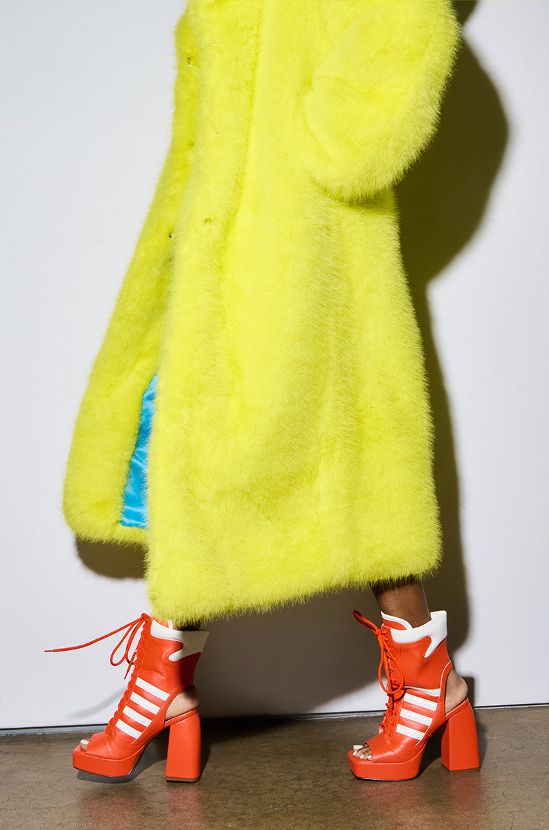 A person dressed in a bright yellow fuzzy coat walks on a concrete floor, their lower half visible against a plain background, showcasing the AZALEA WANG PAMINA ORANGE CHUNKY SANDAL with white stripes and a comfortable cushion insole.