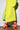 A person dressed in a bright yellow fuzzy coat walks on a concrete floor, their lower half visible against a plain background, showcasing the AZALEA WANG PAMINA ORANGE CHUNKY SANDAL with white stripes and a comfortable cushion insole.