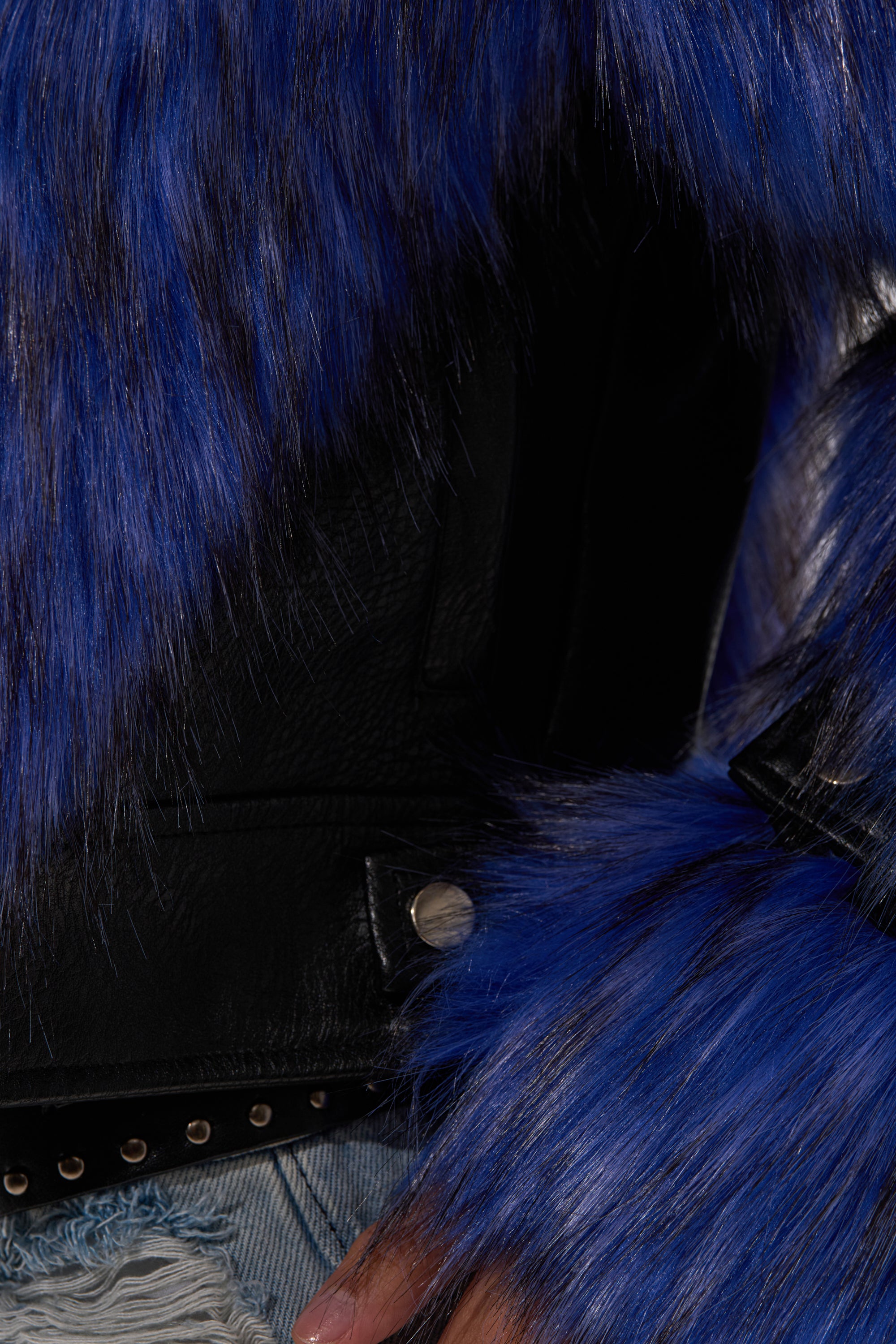 A close-up of a person wearing the ASTRO FUR PANEL MOTO, an electric blue faux fur moto jacket with long, shaggy blue and purple sleeves, silver button accents, paired with light blue ripped denim jeans. 10