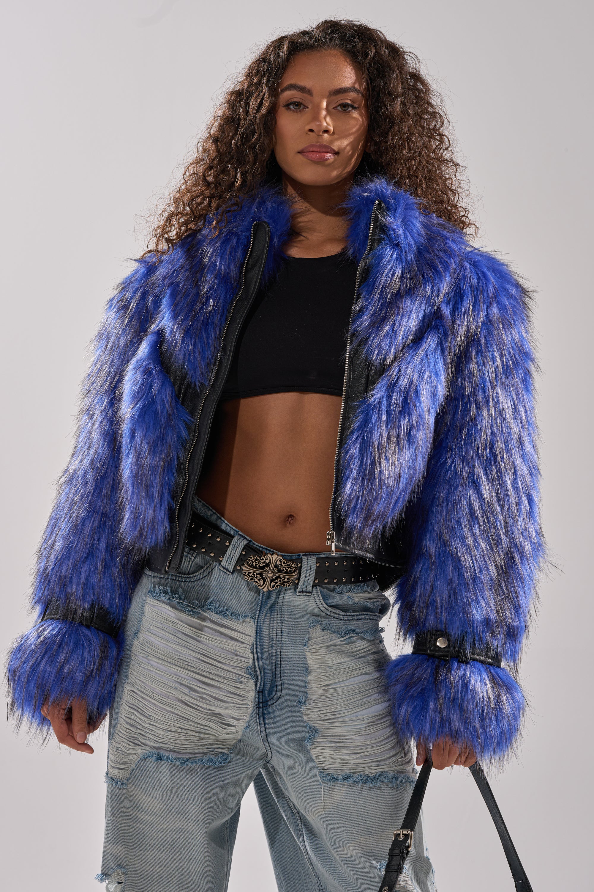 A woman with long curly hair wears the ASTRO FUR PANEL MOTO jacket in electric blue over a black crop top and distressed light-wash jeans with a studded belt, standing confidently and looking at the camera. 1