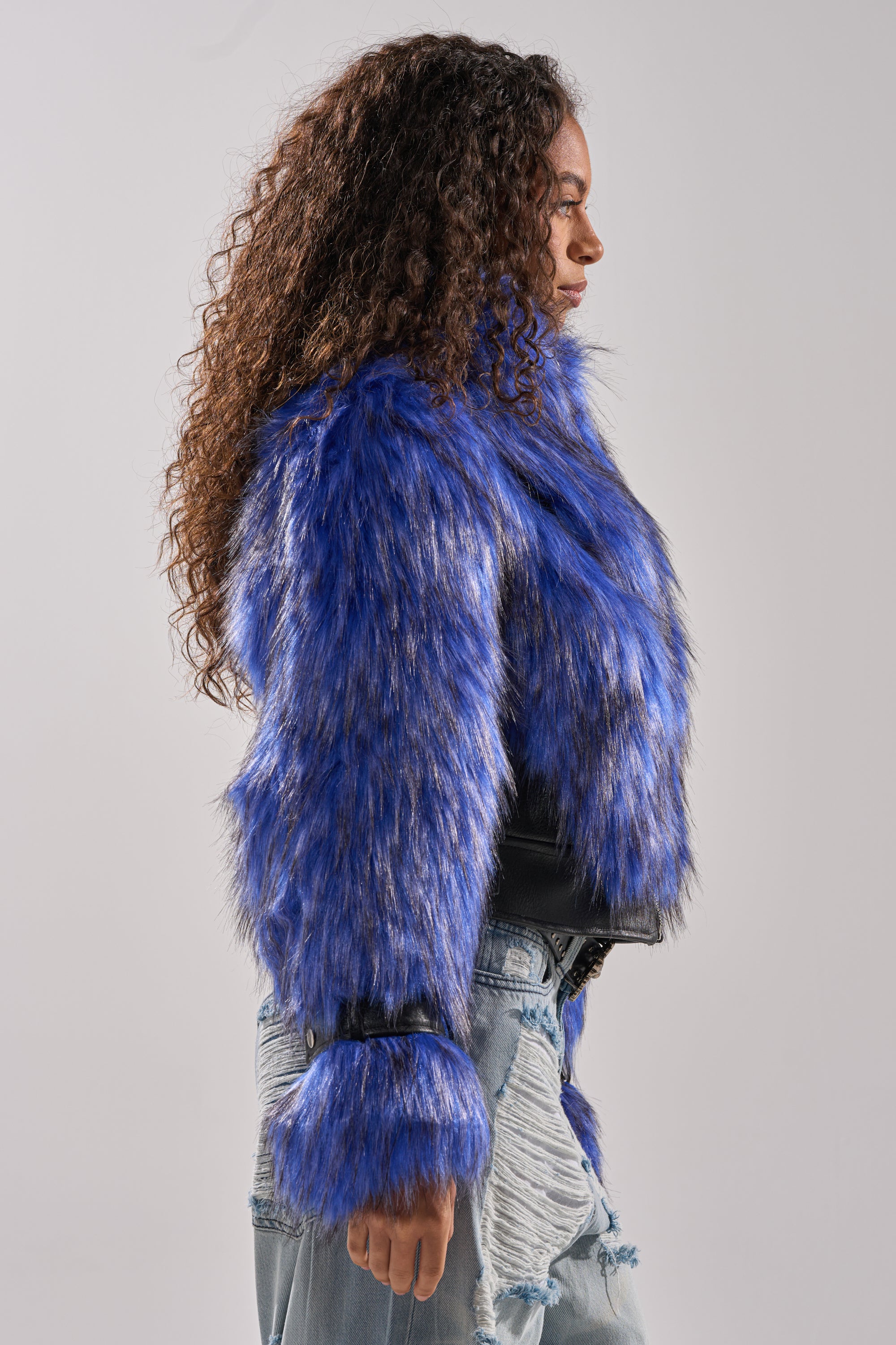 A woman with long curly hair wears the ASTRO FUR PANEL MOTO jacket in electric blue and distressed denim jeans, standing in profile against a plain light gray background. 5