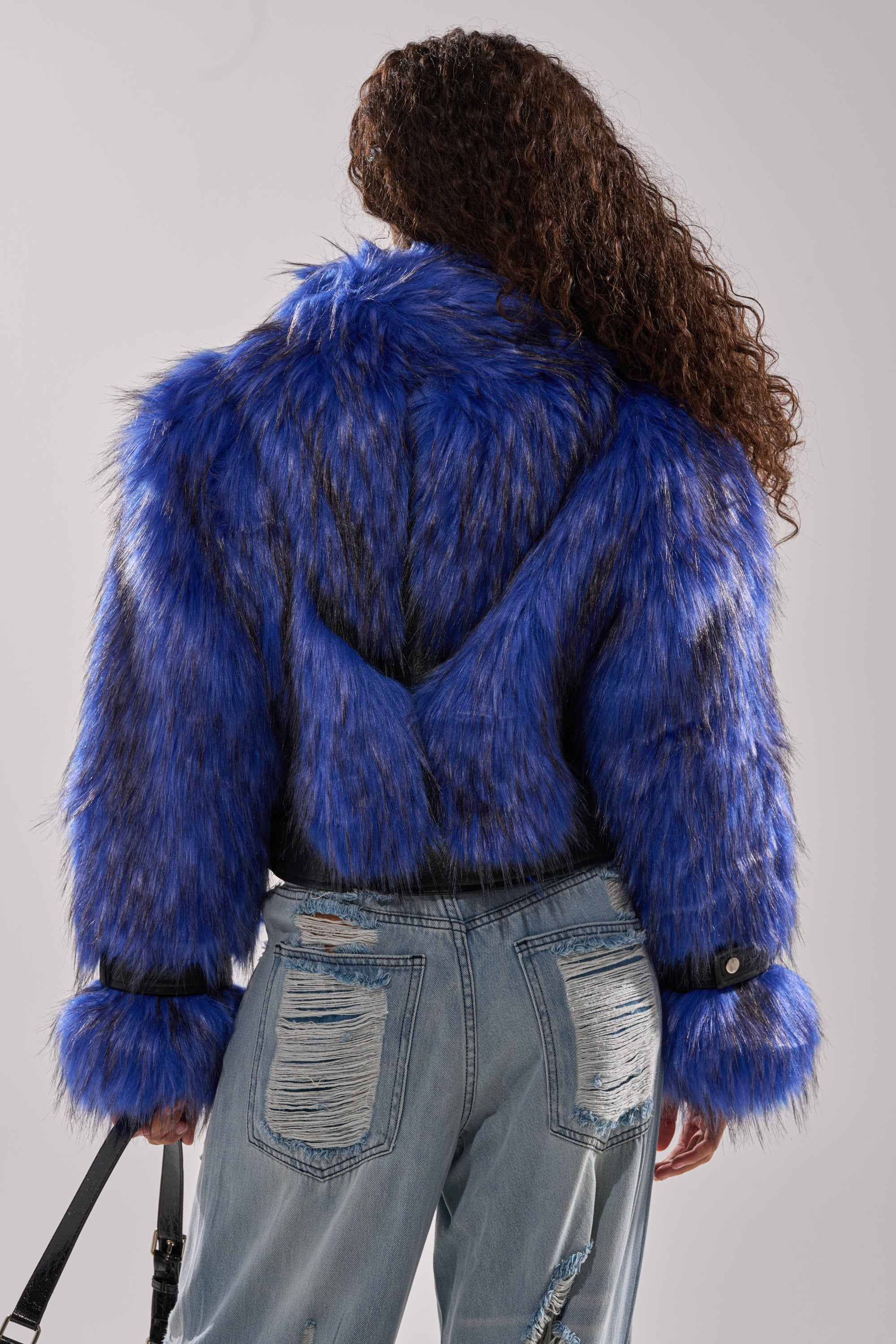 A person with long curly hair wears the ASTRO FUR PANEL MOTO jacket in electric blue and distressed light-wash jeans. The image, taken from behind, showcases the jacket's plush texture and the jeans' rips. 6