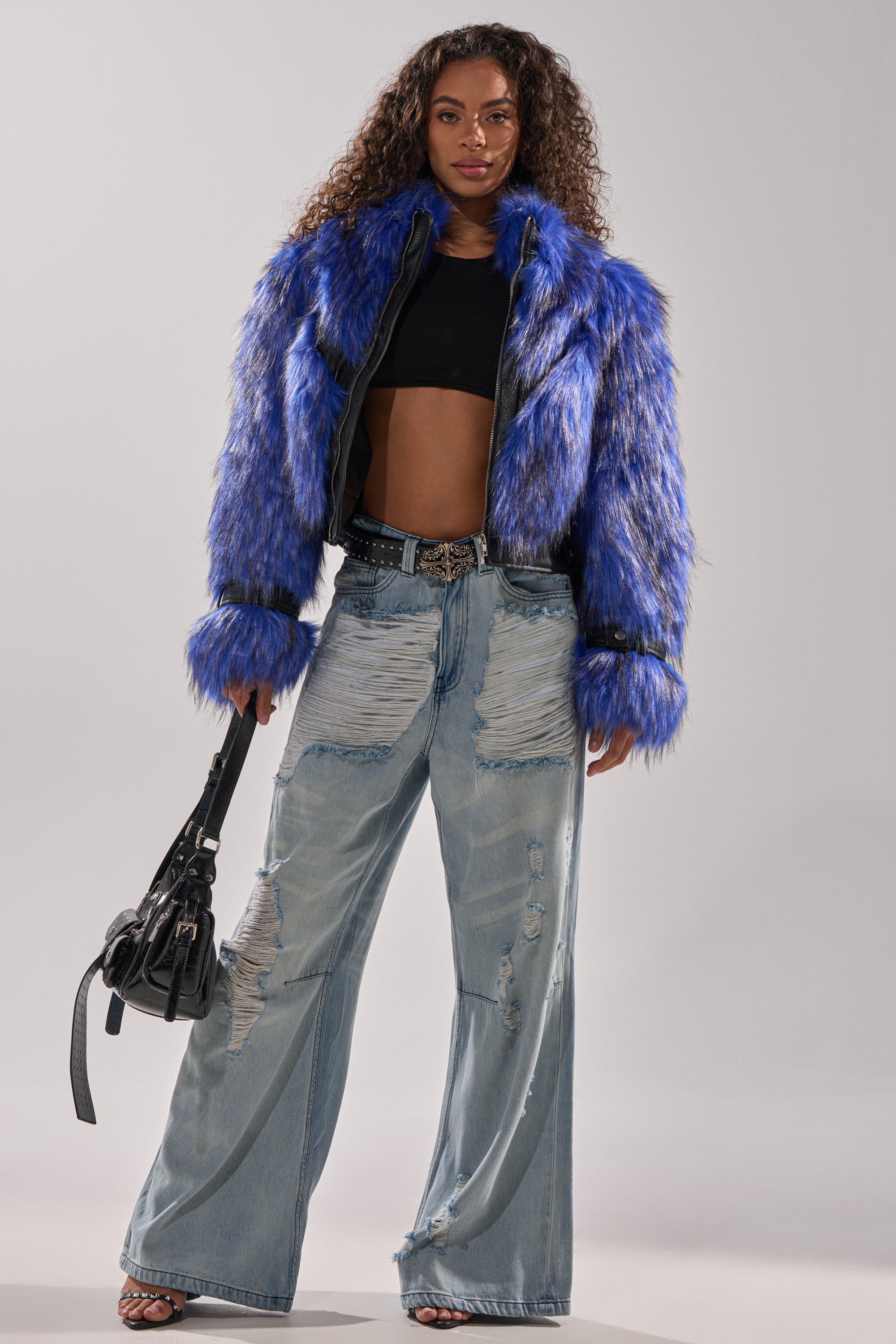 A woman stands against a plain light background wearing the ASTRO FUR PANEL MOTO jacket in electric blue, a black crop top, distressed wide-leg jeans, heeled sandals, and holding a black handbag. 7