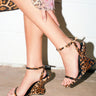 Close-up of a person wearing the AZALEA WANG PANTHERESS LEOPARD WEDGE SANDAL with a suede upper, crossing their legs. A leopard print skirt and sheer floral overlay are visible against a light background.