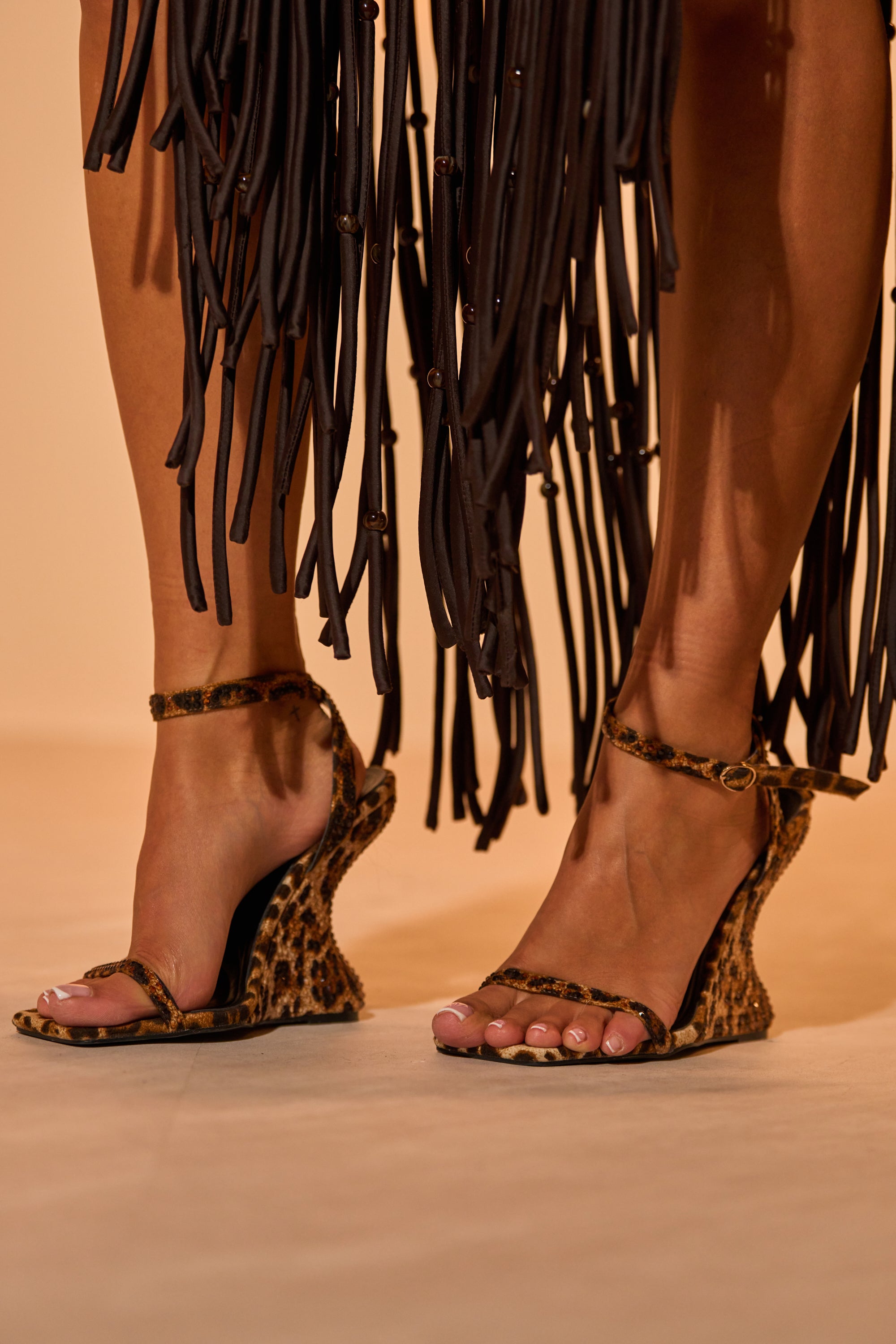 Close-up of a person wearing the AZALEA WANG PANTHERESS LEOPARD WEDGE SANDAL with a black fringe skirt. The suede wedge heels stand out under the fringe, while the light-colored floor highlights the bold leopard design. 1