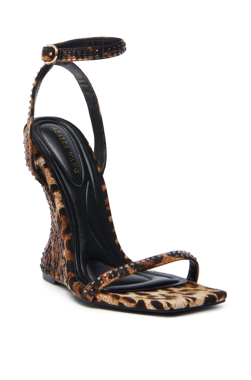 The AZALEA WANG PANTHERESS LEOPARD WEDGE SANDAL features a suede textile upper with an ankle strap, rhinestone embellishments, a unique sculpted wedge heel, and a sleek black insole.