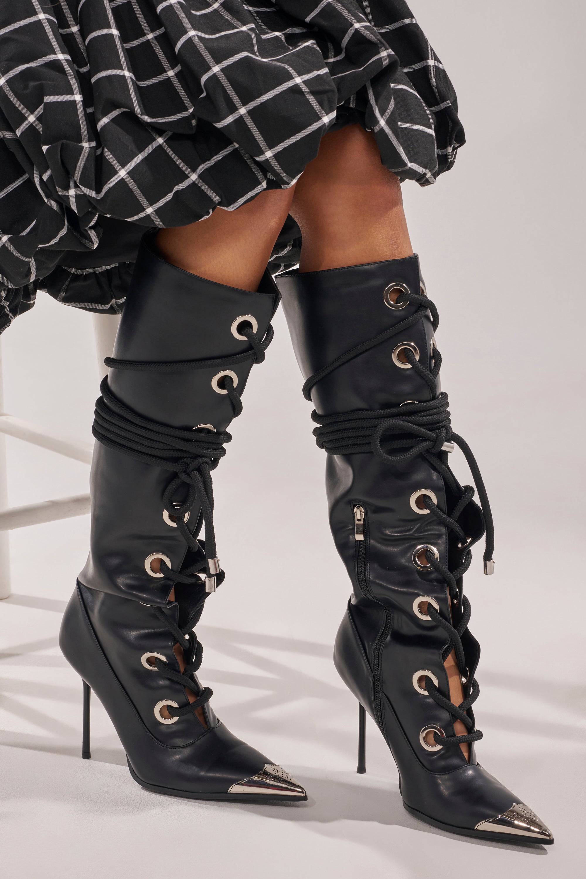 A person wearing the AKIRA GOLD PANTOUM BLACK PU HARDWARE BOOT—black faux leather, knee-high stilettos with metallic pointed toes—paired with a black-and-white checkered skirt. 0