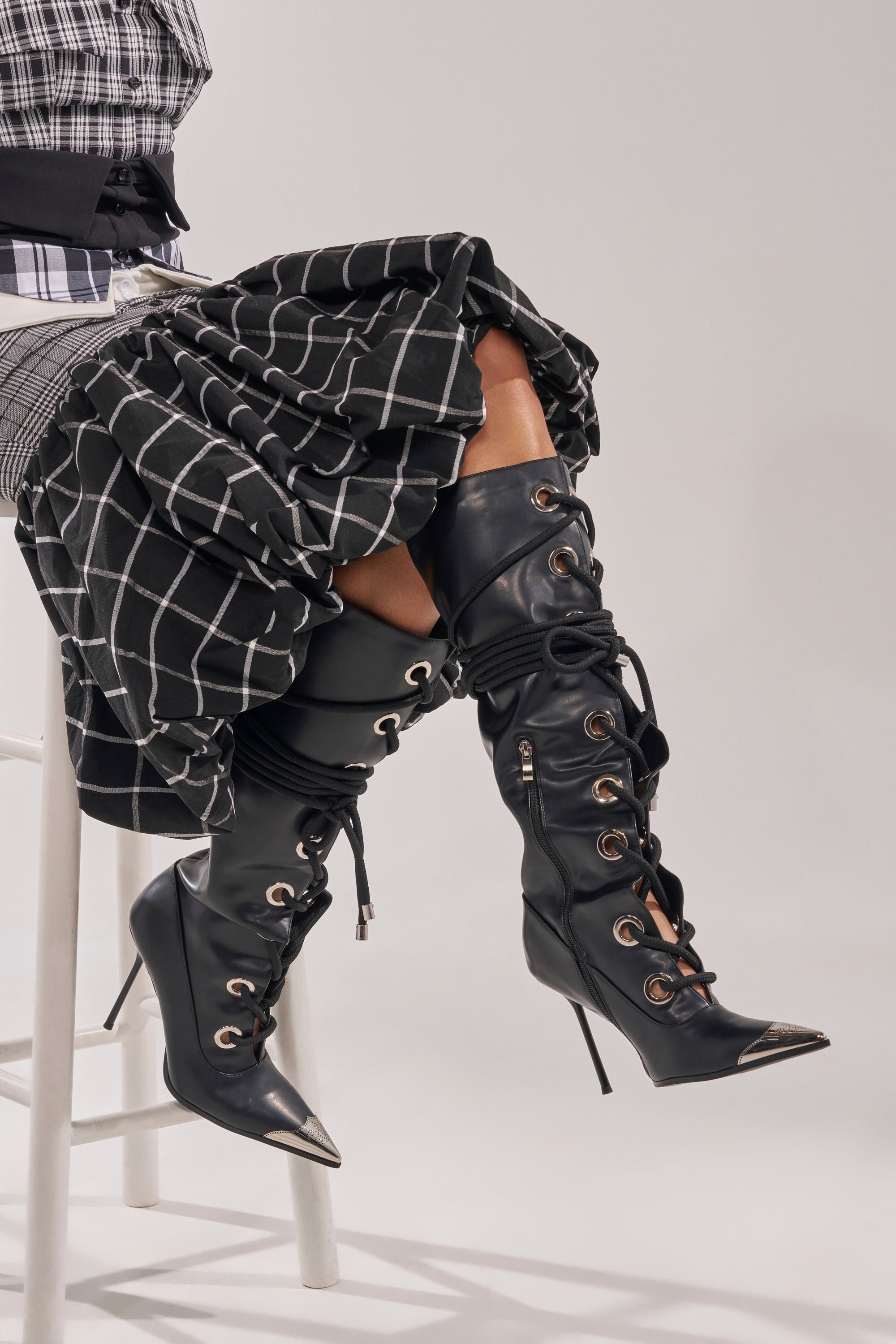 A person sits on a white stool, wearing a black and white plaid skirt with the AKIRA GOLD PANTOUM BLACK PU HARDWARE BOOT—sleek faux leather knee-high boots featuring lace-up fronts and pointed toes. 1
