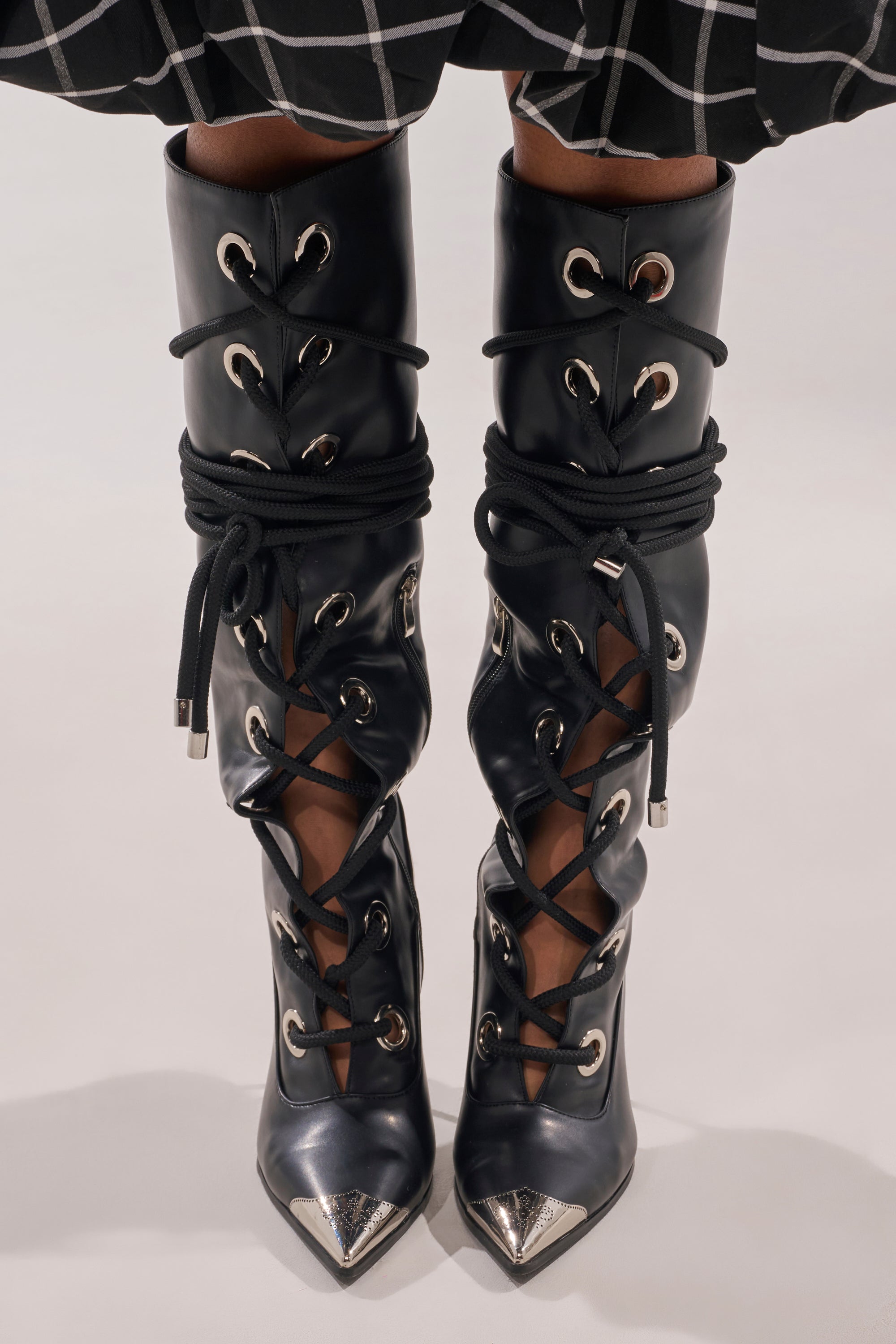 Close-up of the AKIRA GOLD PANTOUM BLACK PU HARDWARE BOOT—black faux leather knee-high stilettos with metallic pointed toes, large silver eyelets, and criss-cross black laces; a checkered skirt appears just above. 3