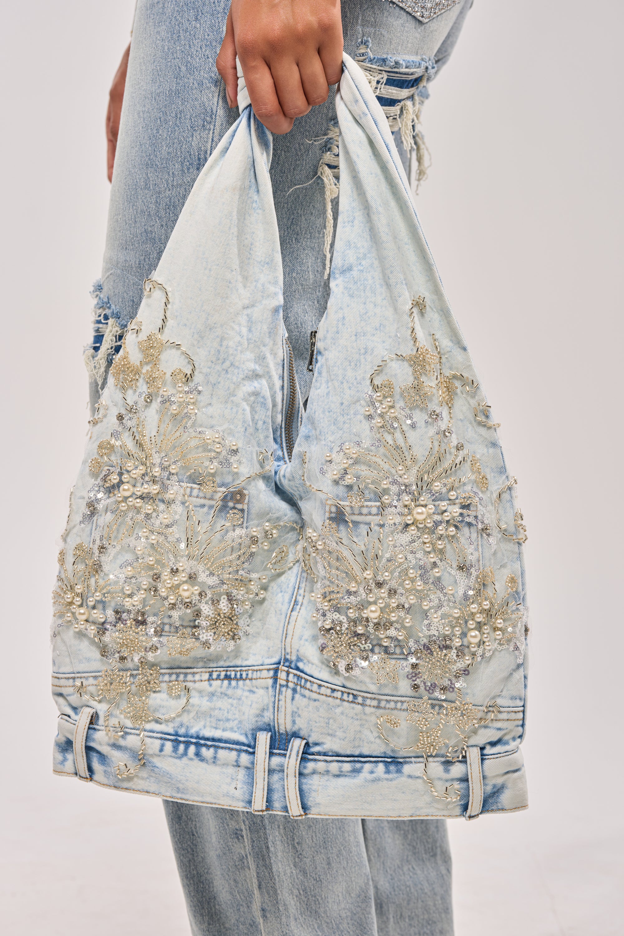 A hand holds the BLESSING IN DENIM EMBELLISHED SHOULDER BAG—light wash denim with silver embroidery and sequins—paired with distressed light blue jeans in a matching faded finish. 1