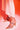 Someone wears white pants and the AZALEA WANG PAOLINA GOLD SANDAL, featuring clear high heels with large sparkling rhinestones, while standing on a surface lit in pink and orange.