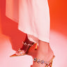 Someone wears white pants and the AZALEA WANG PAOLINA GOLD SANDAL, featuring clear high heels with large sparkling rhinestones, while standing on a surface lit in pink and orange.