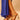 A person in a royal blue dress and AZALEA WANG PAOLINA GOLD SANDALS with jeweled accents stands on a beige surface.