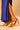 A person in a royal blue dress and AZALEA WANG PAOLINA GOLD SANDALS with jeweled accents stands on a beige surface.