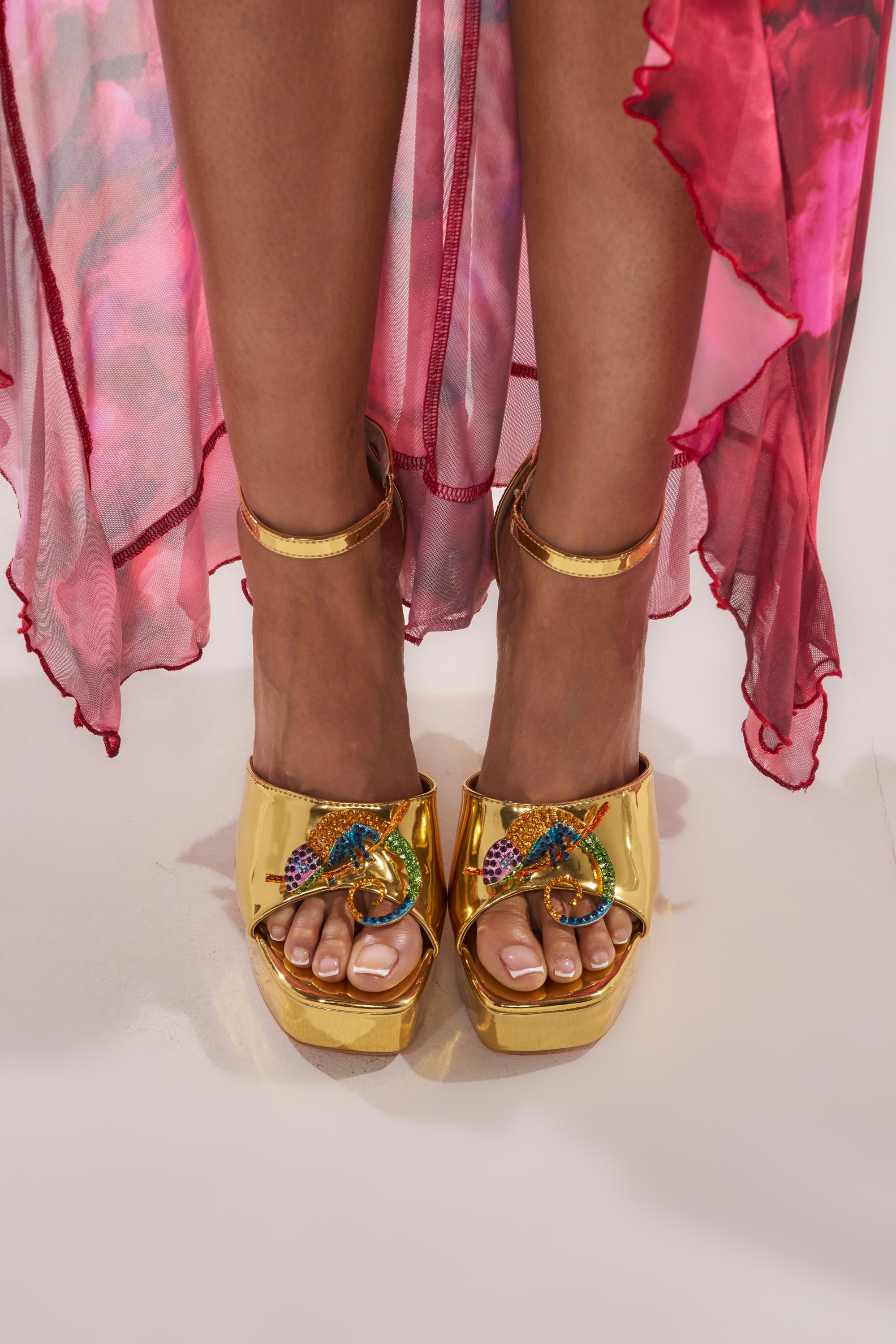 A person stands on a light surface wearing the AZALEA WANG PASQUALE GOLD IGUANA EMBELLISHED SANDAL, paired with a pink and red sheer, flowy dress draped around their legs. 0