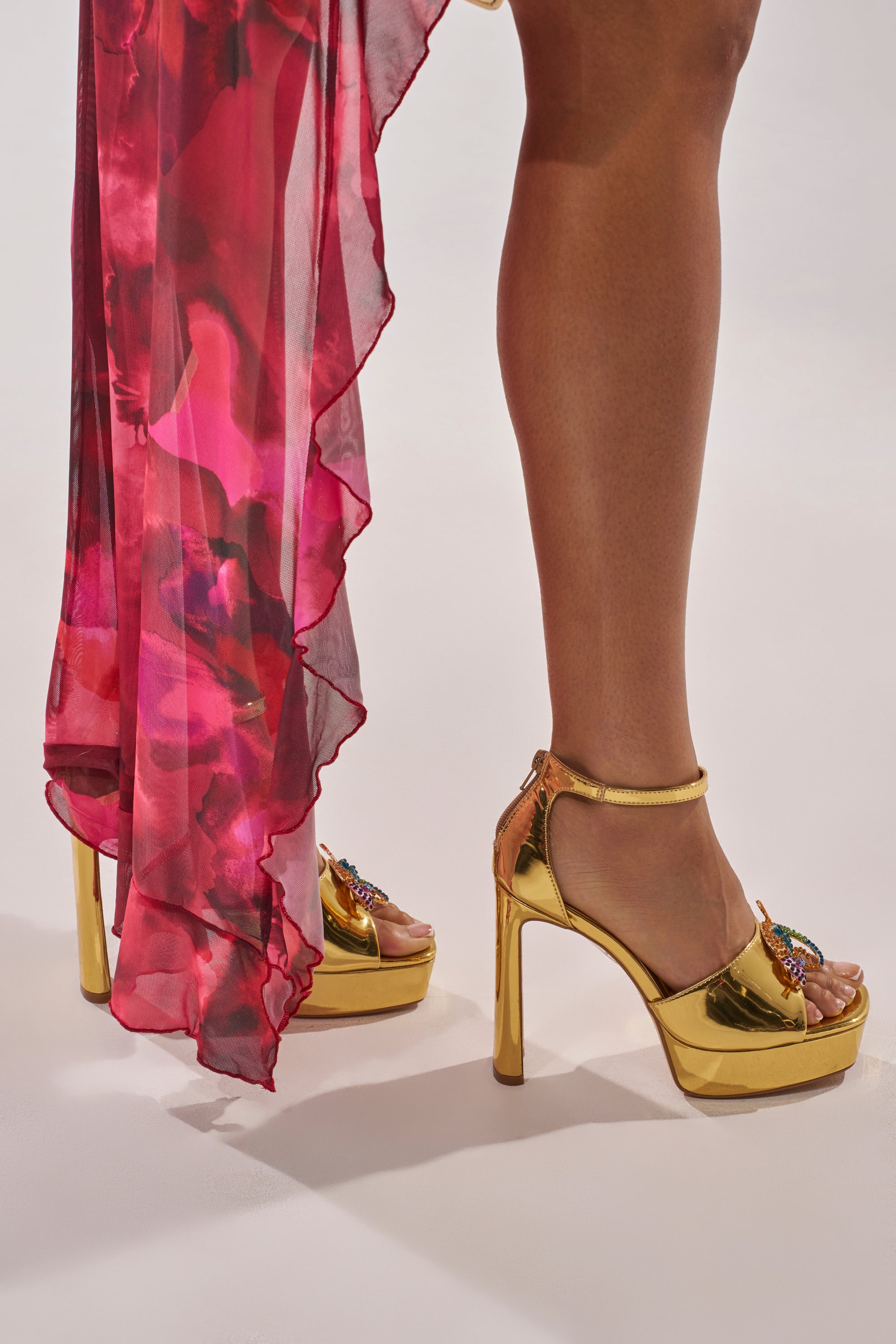 A person wearing a flowing, sheer pink floral dress and AZALEA WANG PASQUALE GOLD IGUANA EMBELLISHED SANDALS stands on a white surface. 11