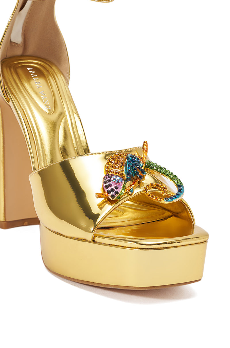 A close-up of the AZALEA WANG PASQUALE GOLD IGUANA EMBELLISHED SANDAL—a shiny gold faux leather platform heel featuring a gold, green, blue, and purple snake-shaped ornament on the front strap. 10
