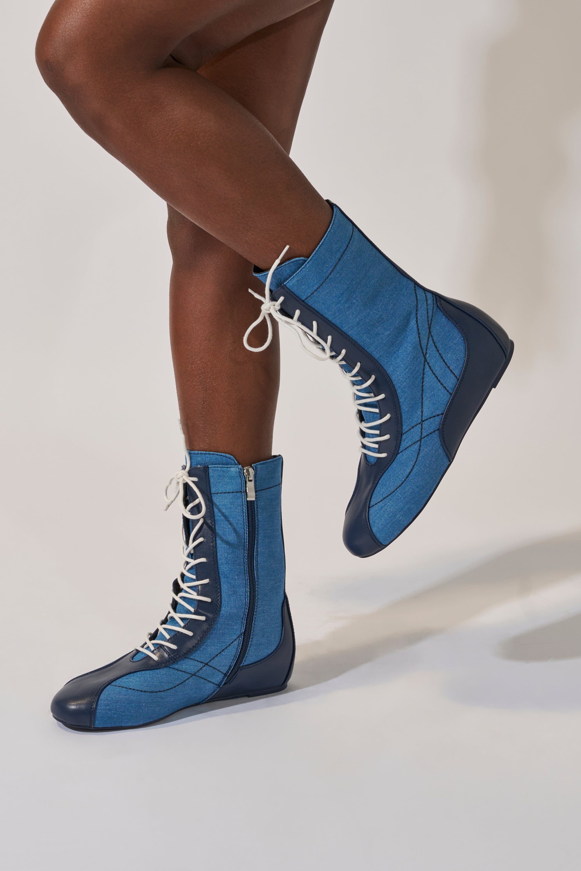 Someone models the AZALEA WANG PASSER NAVY FLAT SNEAKER, a mid-calf navy denim shoe featuring white laces and black soles, standing on one leg with the other bent against a plain light background.