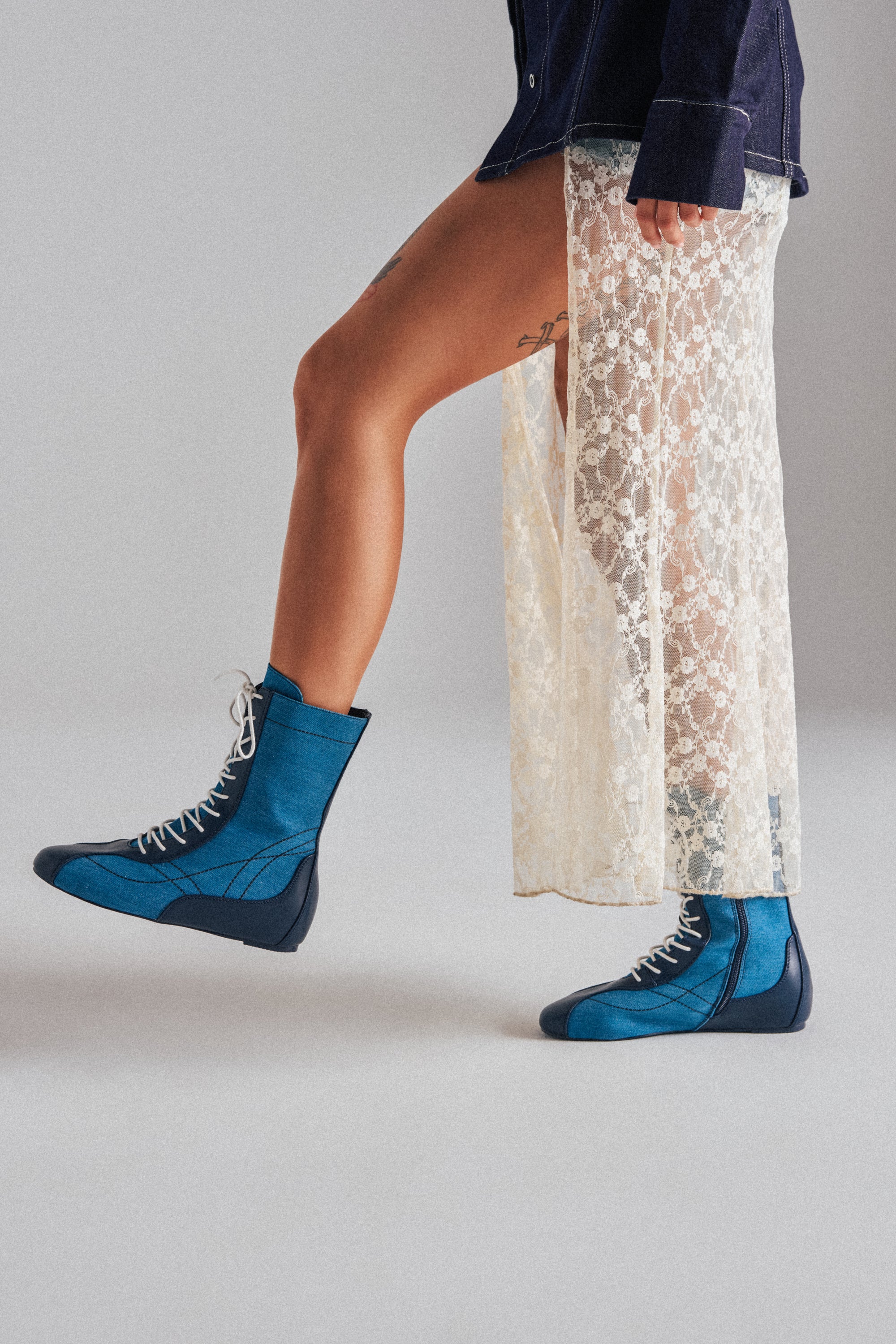 Wearing AZALEA WANG PASSER NAVY FLAT SNEAKERS, a denim jacket, and a sheer cream lace skirt that reveals a thigh tattoo, a person stands against a plain gray background.