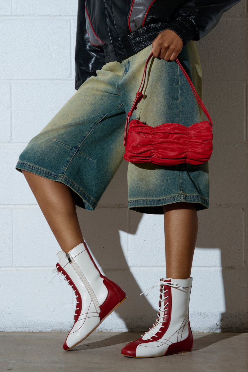 A person stands against a white wall, wearing a black jacket, faded knee-length denim shorts, and AZALEA WANG PASSER sporty flat booties with lace-up detailing. They hold a small red handbag that matches the red accents on their boots.