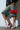 A person stands against a white wall, wearing a black jacket, faded knee-length denim shorts, and AZALEA WANG PASSER sporty flat booties with lace-up detailing. They hold a small red handbag that matches the red accents on their boots.
