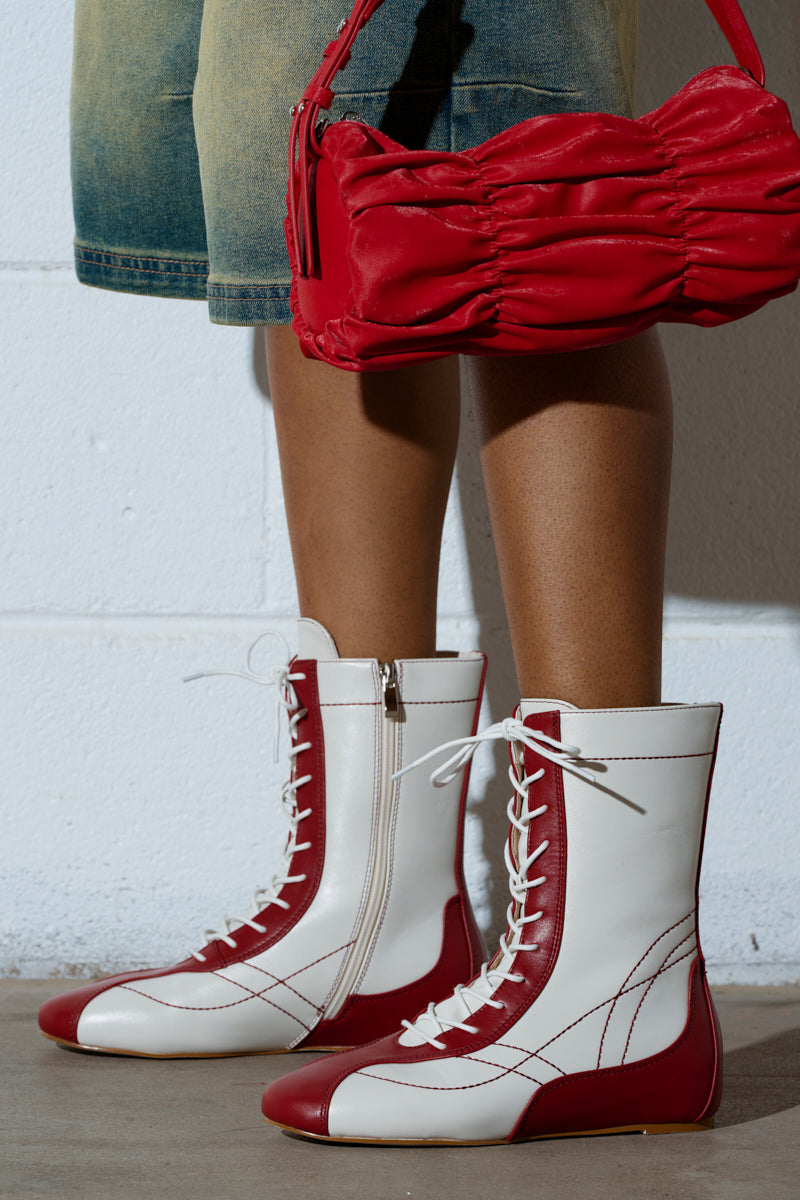 Wearing AZALEA WANG PASSER SPORTY FLAT BOOTIE in white and red with laces and side zippers, a person stands on concrete holding a red ruched handbag. Against a white wall backdrop, they sport shorts that exude casual flair.