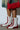 Wearing AZALEA WANG PASSER SPORTY FLAT BOOTIE in white and red with laces and side zippers, a person stands on concrete holding a red ruched handbag. Against a white wall backdrop, they sport shorts that exude casual flair.