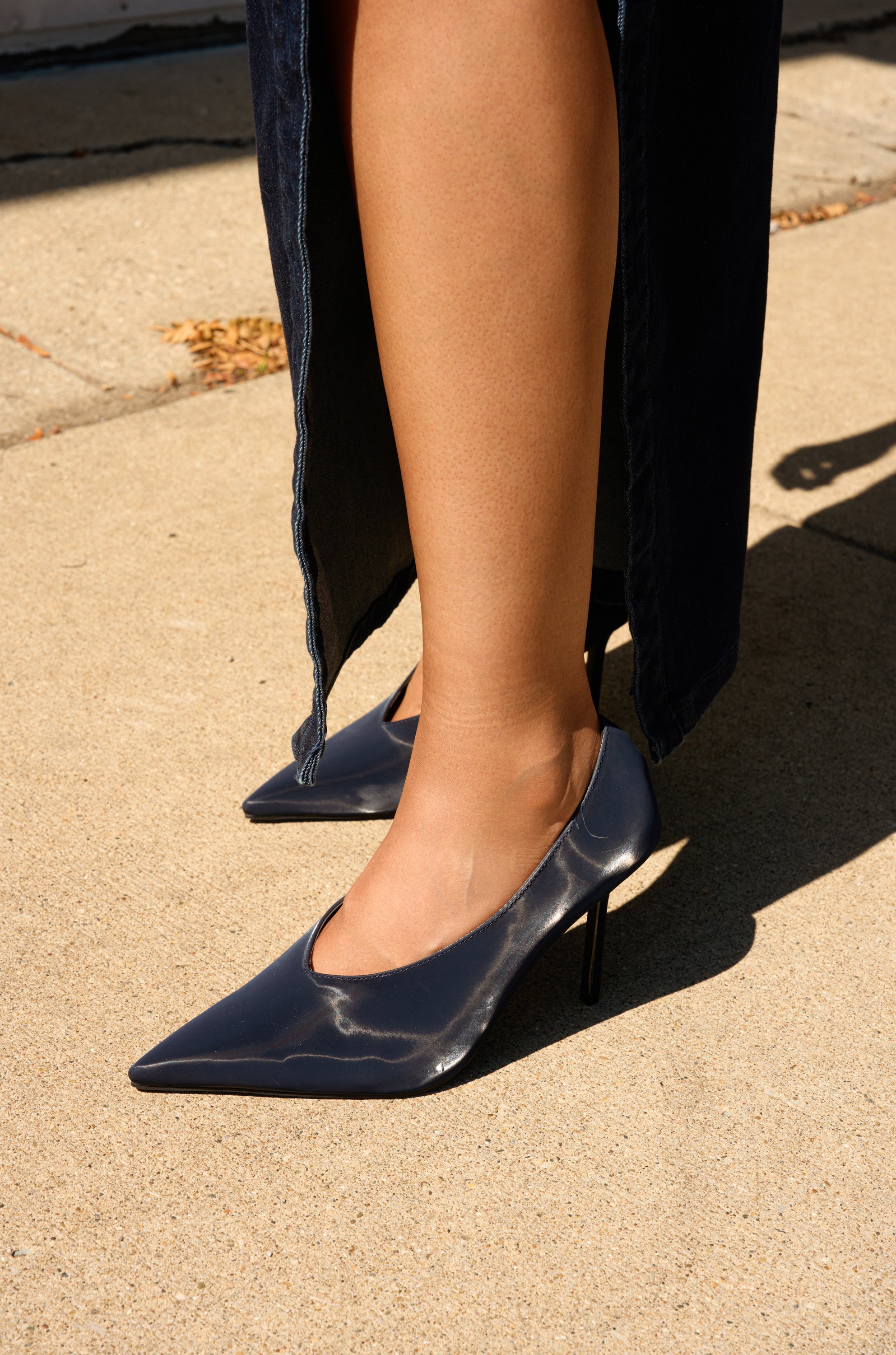 A person stands on a sunlit concrete sidewalk wearing a long dark skirt and the AZALEA WANG PATRICC NAVY LUXE MATERIALS PUMP with a pointed toe and stiletto heel.