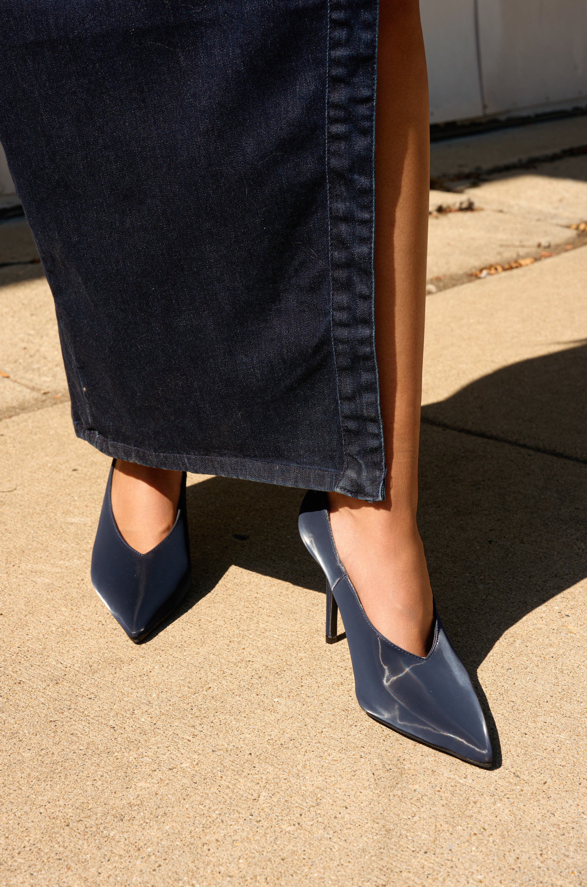 A person stands on a sunlit sidewalk wearing a dark denim skirt with a slit and the AZALEA WANG PATRICC NAVY LUXE MATERIALS PUMP.