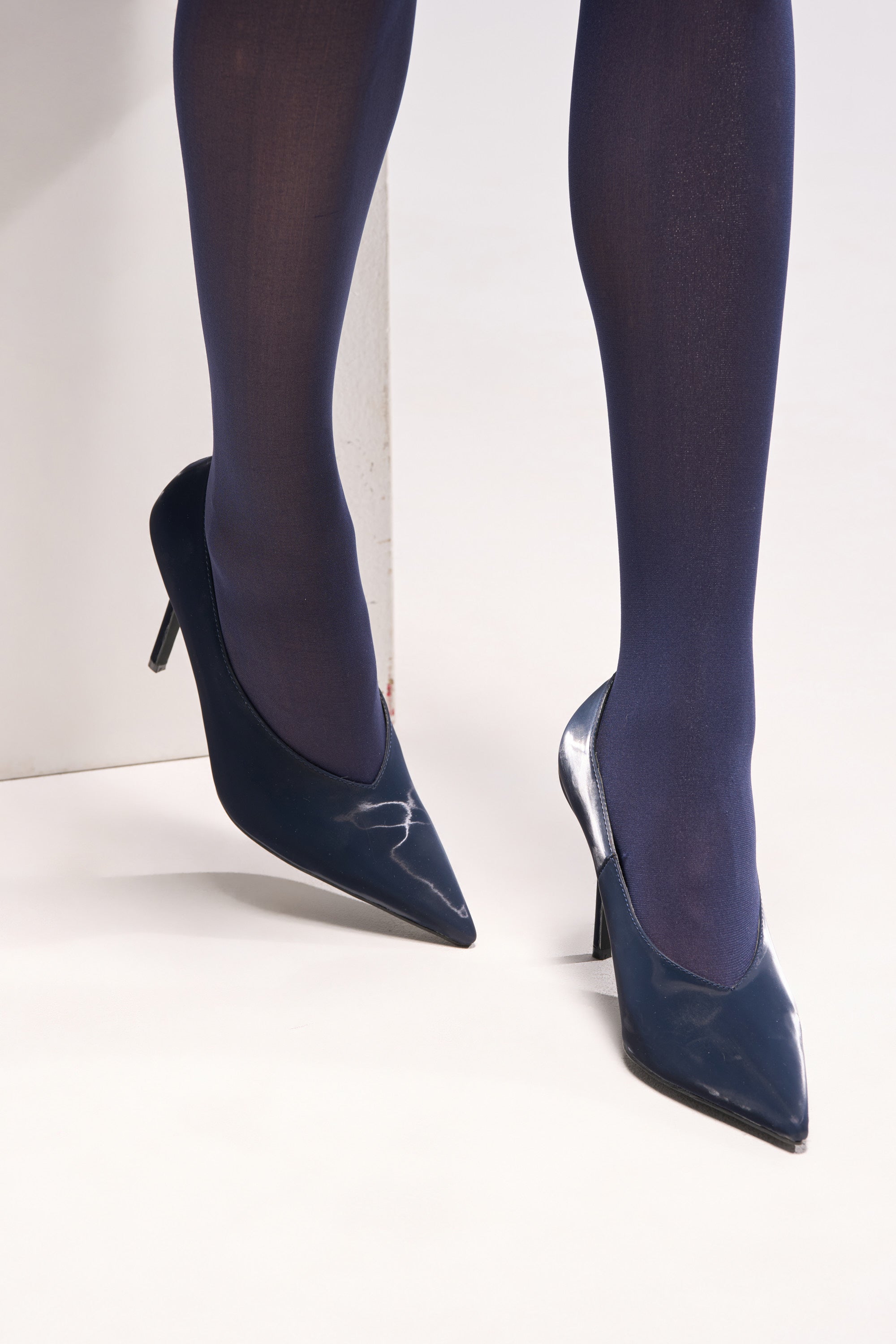 A person wears navy blue tights and the AZALEA WANG PATRICC NAVY LUXE MATERIALS PUMP with stiletto heels, standing against a white background with one foot slightly raised and toes pointed down.