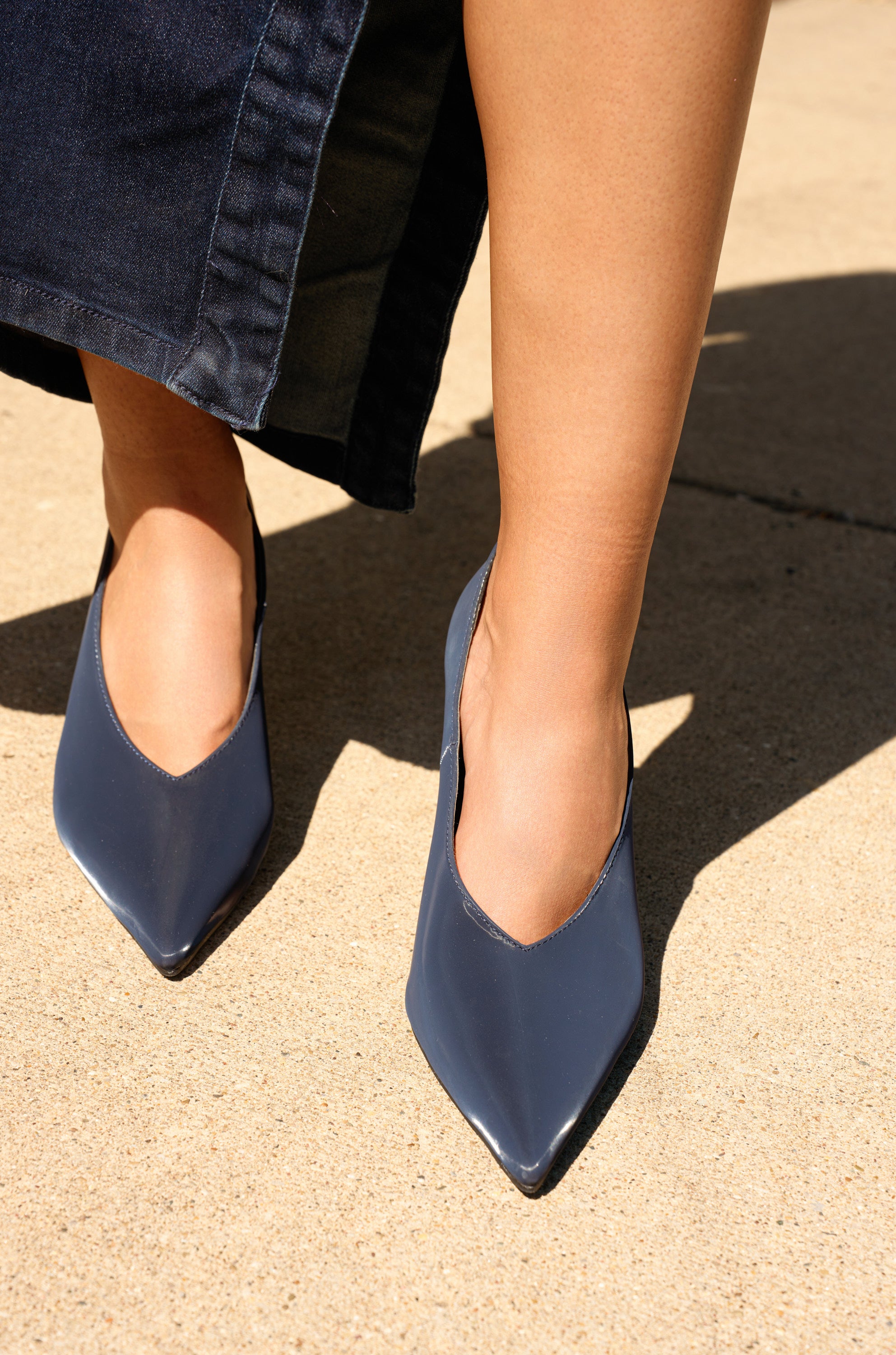 A person stands on a sunlit concrete surface wearing a dark denim skirt and the AZALEA WANG PATRICC Navy Luxe Materials Pump, featuring shiny pointed toes.