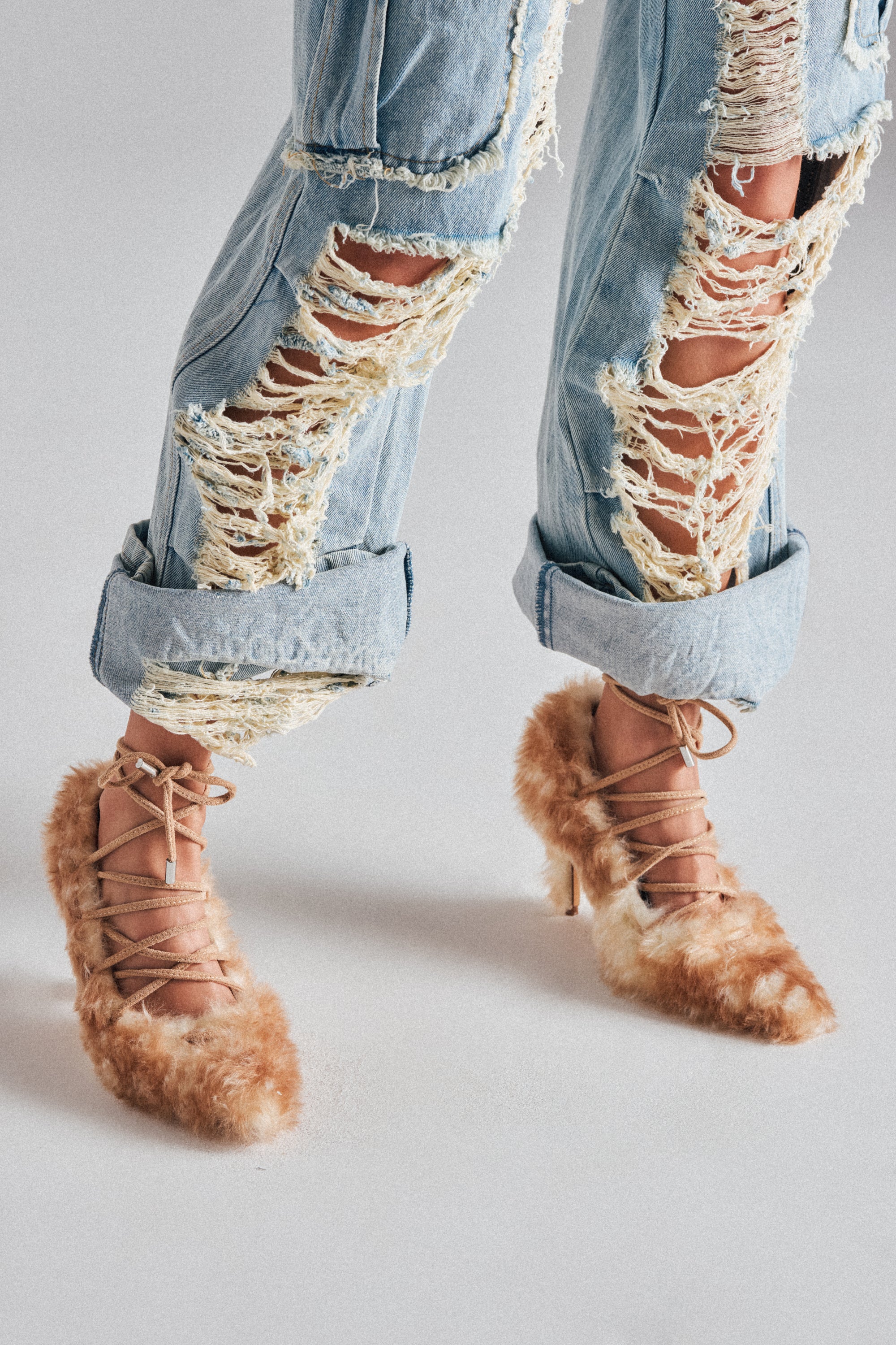 A person stands against a light background wearing ripped, rolled-up blue jeans and AZALEA WANG PAULHINO SNOW LEOPARD STRAPPY PUMP heels featuring tan faux fur and crisscross laces.