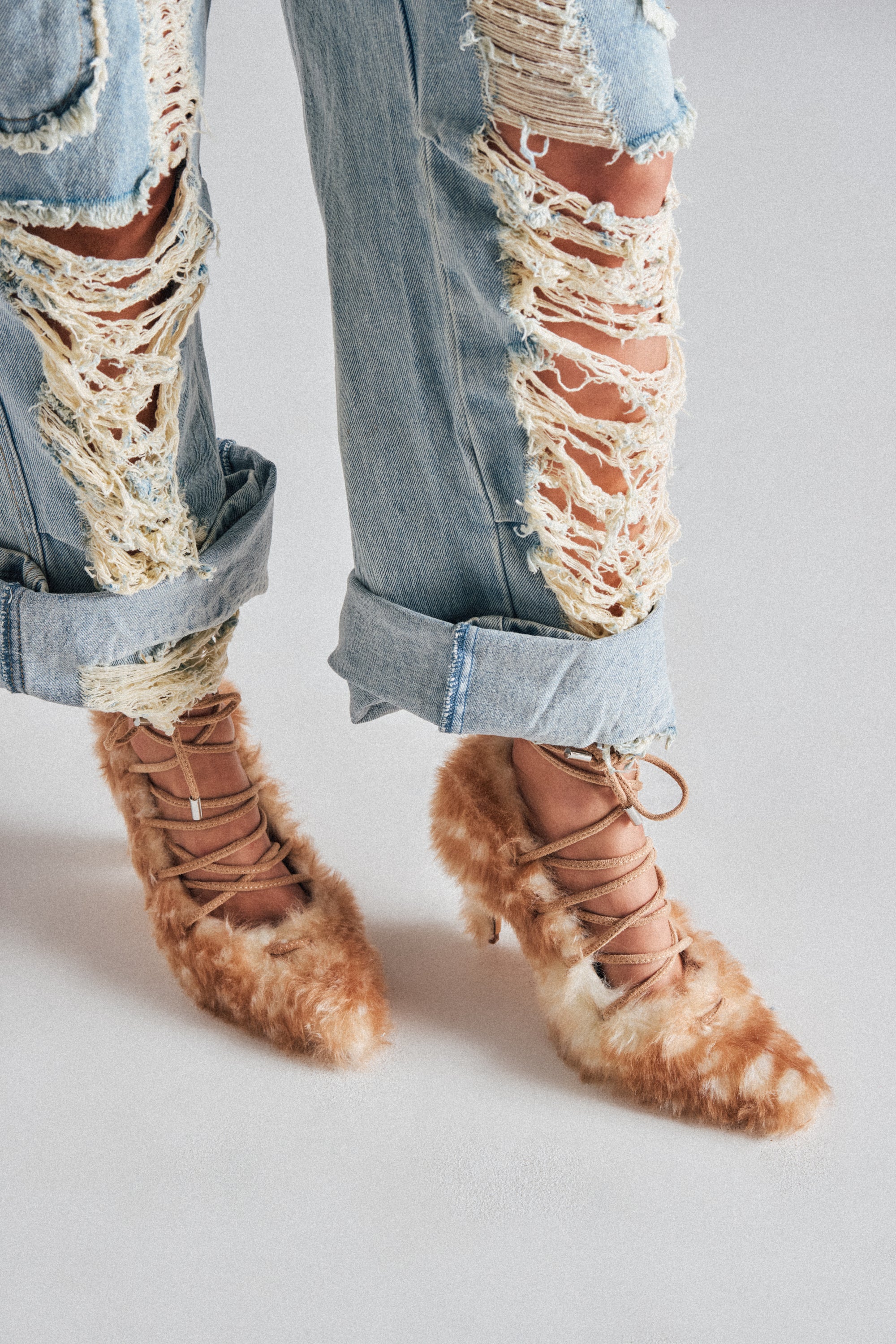 A person wearing light blue, heavily distressed jeans with large rips and AZALEA WANG PAULHINO SNOW LEOPARD STRAPPY PUMPS stands on a light-colored surface.