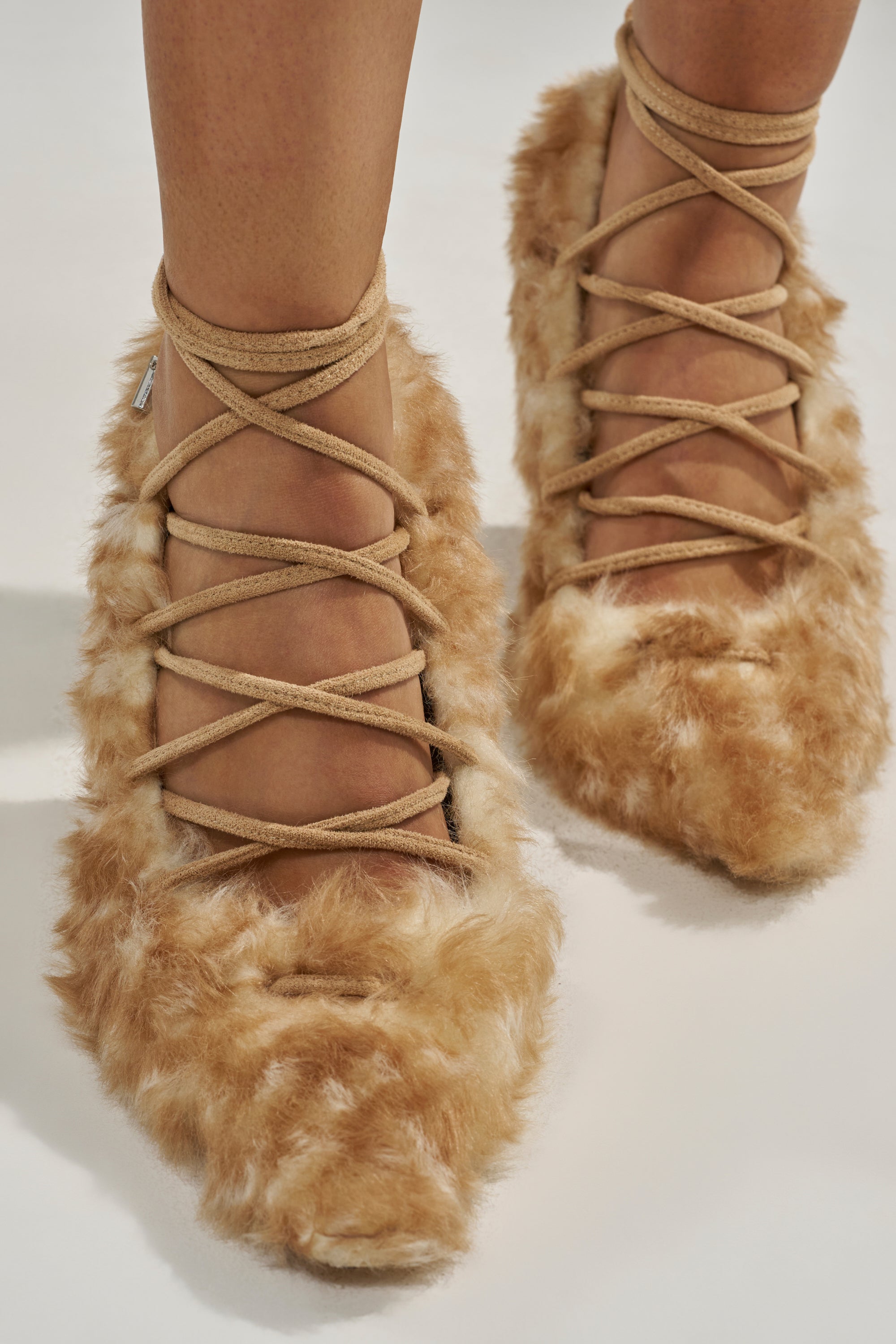 Close-up of a person's feet wearing the AZALEA WANG PAULHINO SNOW LEOPARD STRAPPY PUMP—tan, fluffy heels with pointed toes, crisscrossing suede ankle straps, and faux fur covering the entire foot for a unique, textured look.