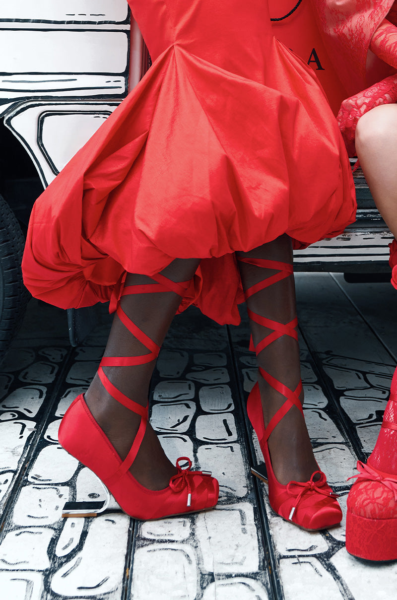 Dressed in sophisticated, bold red attire, two individuals don stunning AZALEA WANG PAVARITI BALLET SWOOP HEELS IN RED. These heels showcase elegant crisscross straps and a sleek satin upper. Against a black and white illustrated backdrop that evokes the charm of a vintage car, they exude an effortlessly chic and fashion-forward aura.