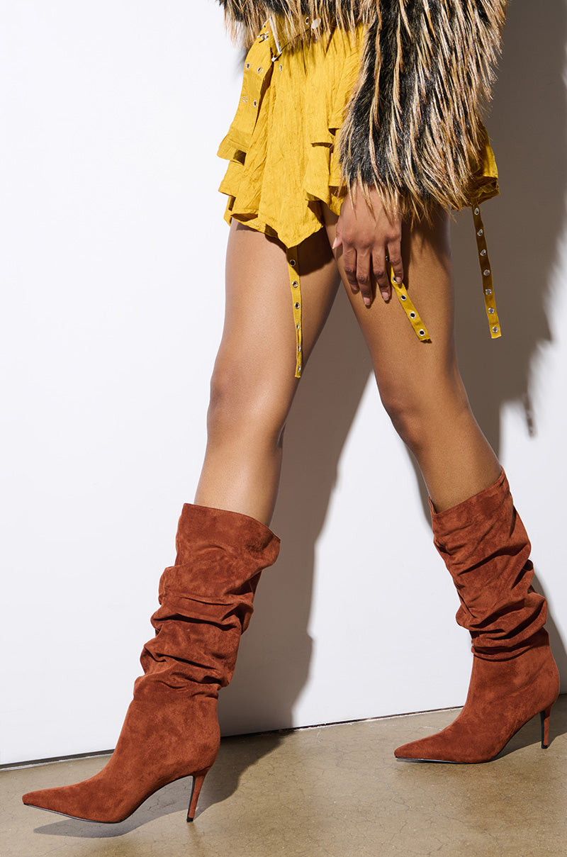A person sporting the AZALEA WANG PAVETTA Brown Faux Suede Slouch Boots and a yellow ruffled skirt strides confidently across a concrete floor in front of a plain white wall, with their fur jacket adding an extra touch of style.