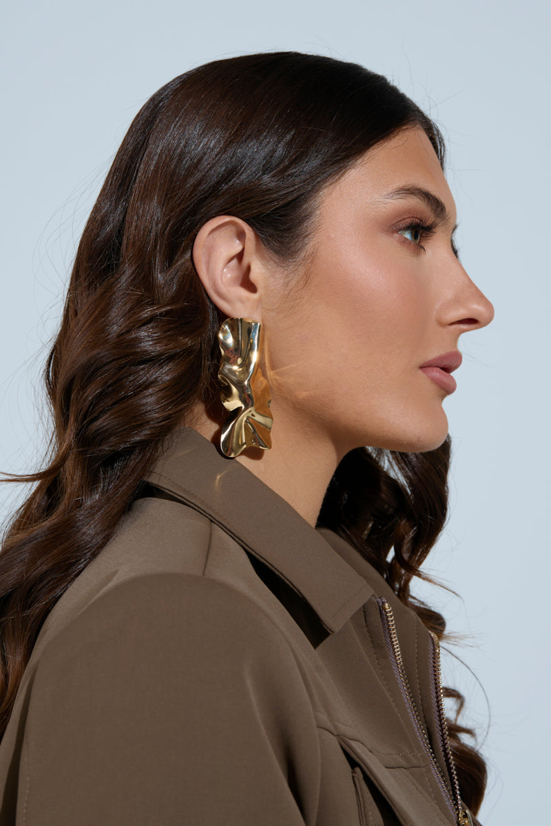 A woman with long brown hair wears a brown jacket and large ITS ORGANIC EARRING with textured pendants. She looks serene, gazing sideways against a light blue background. 1