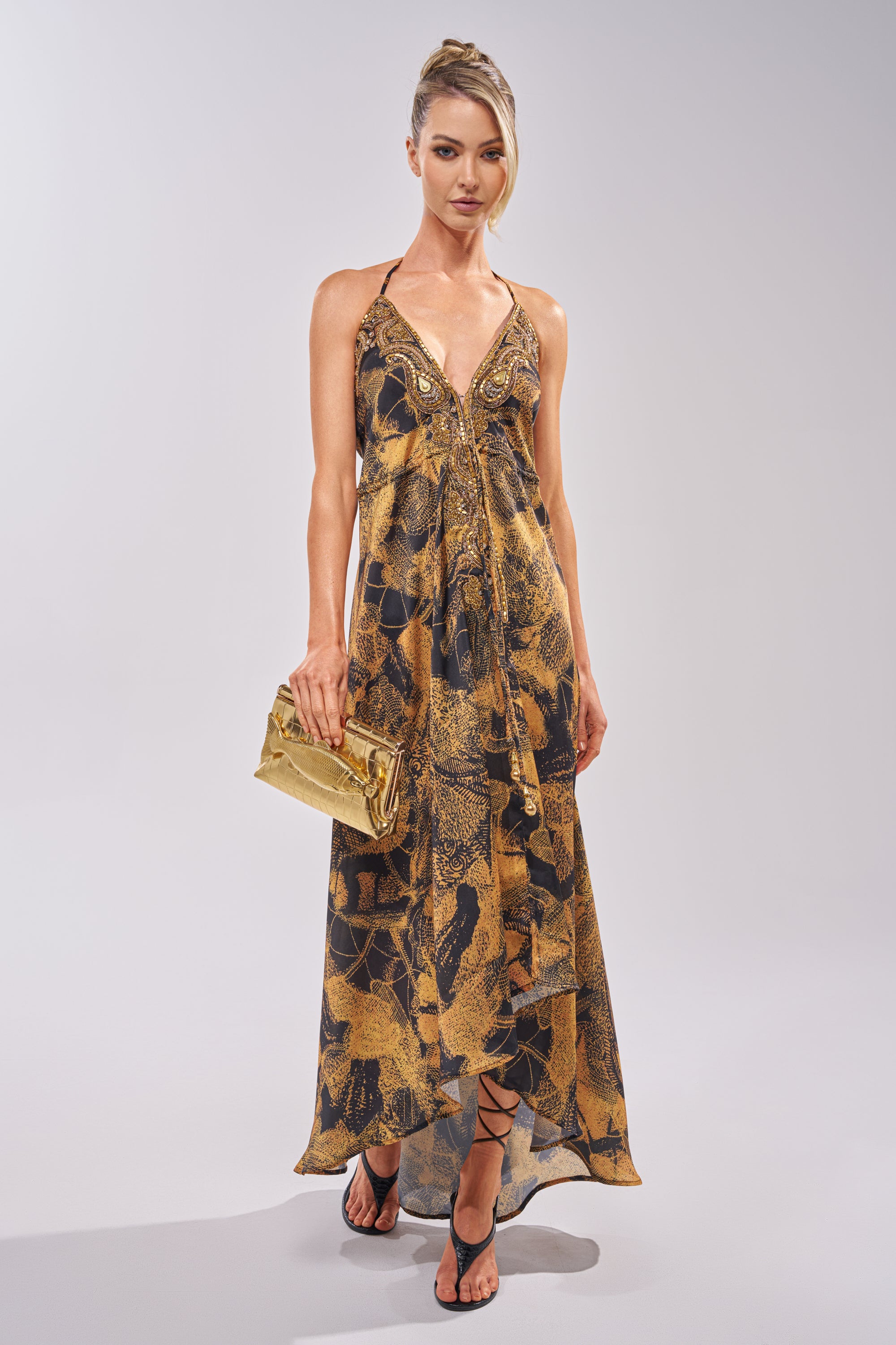 A woman models the SHINE ON YOU PRINTED MAXI DRESS, gold clutch in hand. Her hair is up and she completes the look with black strappy sandals, posing against a plain light background.