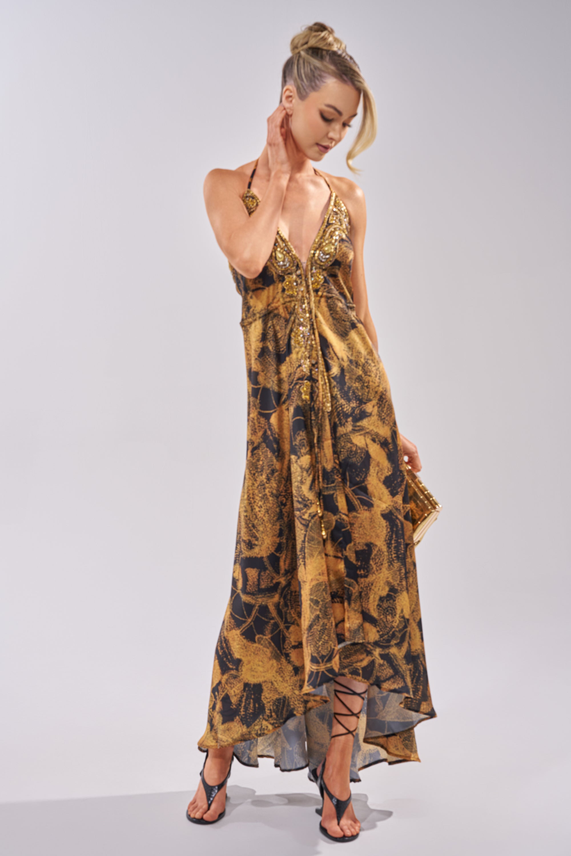 Wearing the SHINE ON YOU PRINTED MAXI DRESS, a woman stands gracefully in black strappy heels, holding a gold clutch, her hair in an elegant updo, and touching her neck.