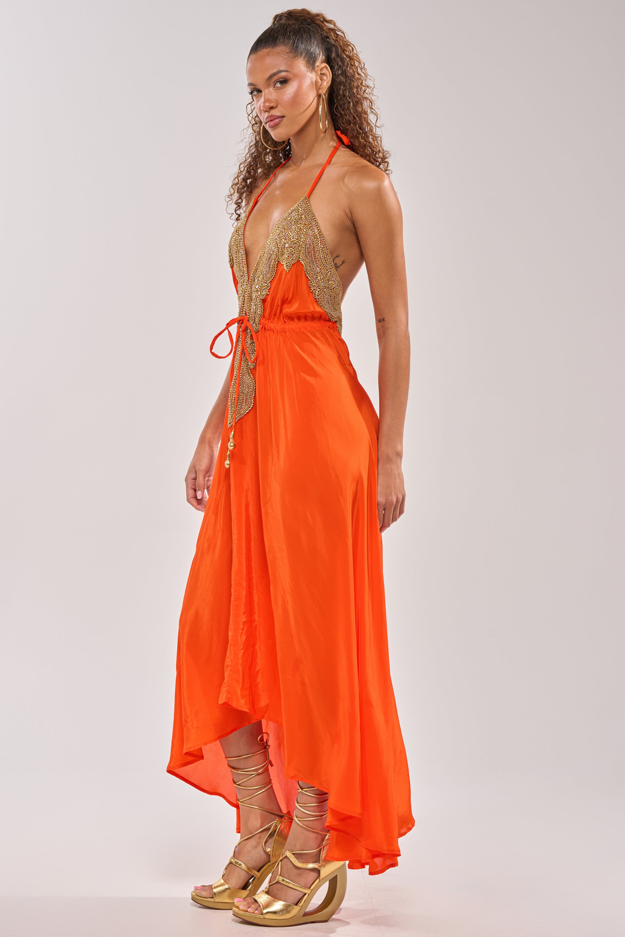 Woman in the SORRY NOT SORRY EMBELLISHED MAXI DRESS with gold lace details, paired with gold strappy heels and curly hair, poses against a light background—ideal for evening occasions.
