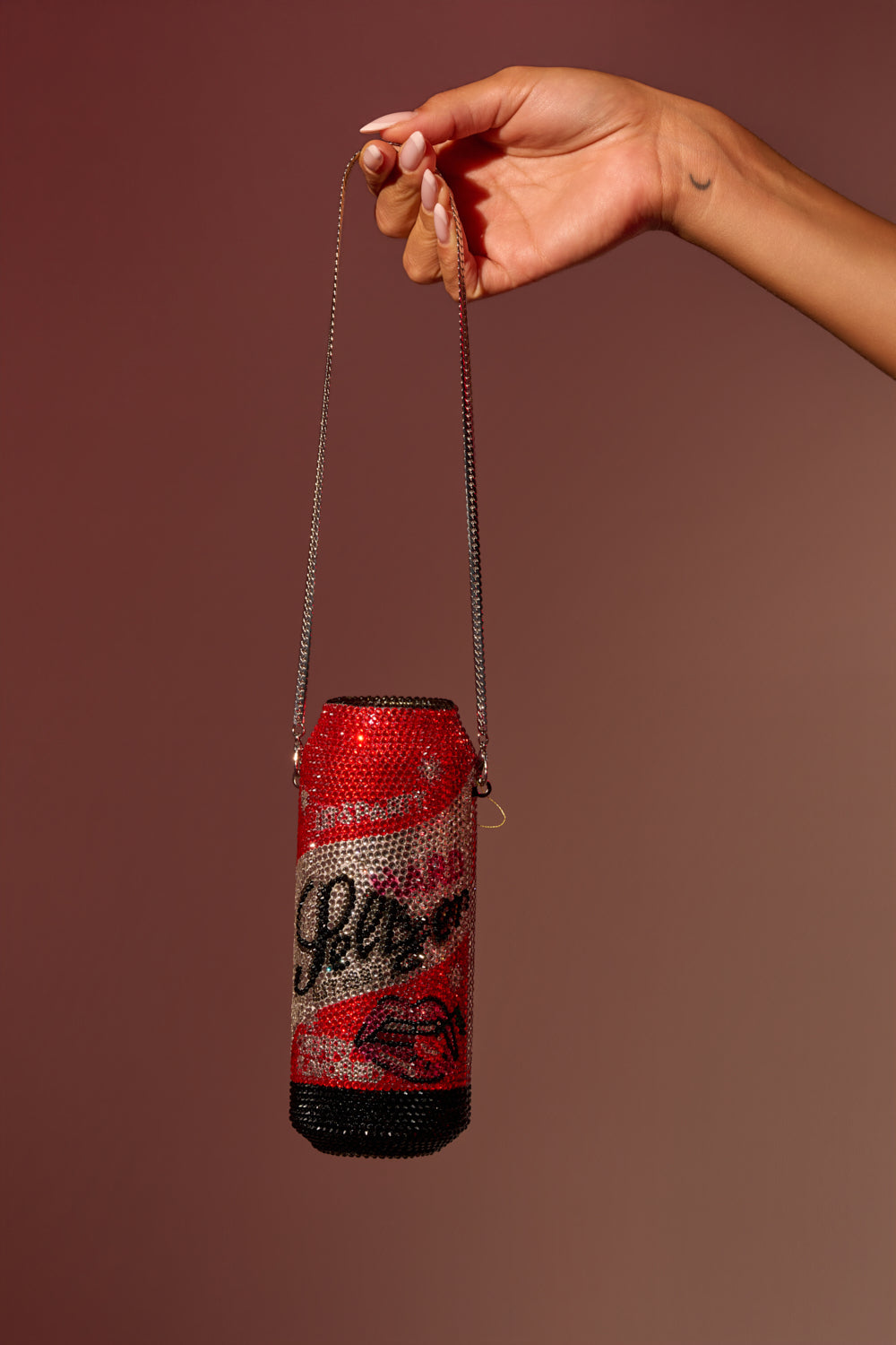 A manicured hand holds the TAKE A SIP BLING BAG, a sparkling rhinestone soda can purse with red, silver, and black script-like designs—a bold street style statement against a brown background. 0
