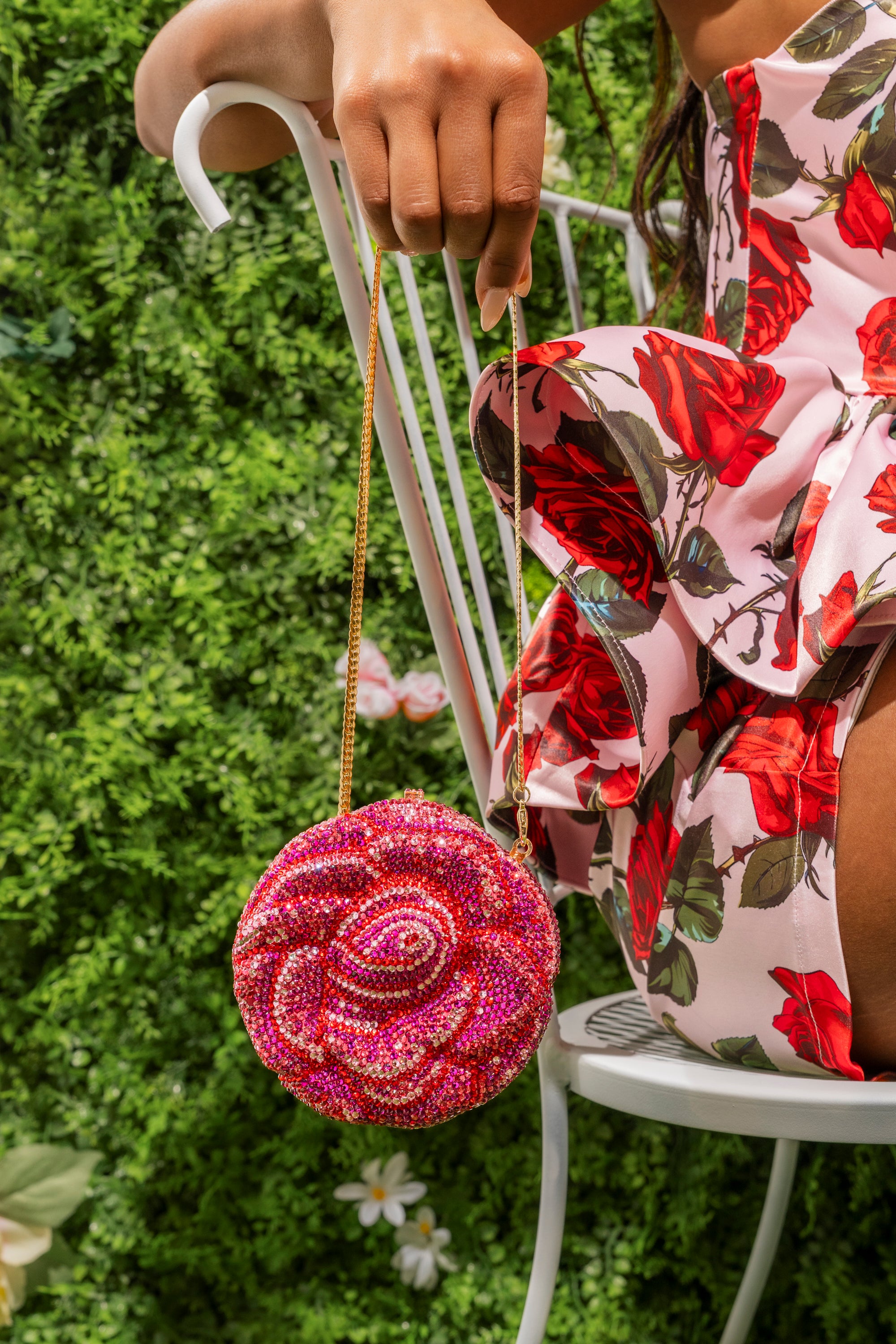 A person in a floral dress sits on a white chair, holding the NOT THAT KIND OF ROSE PURSE—a rhinestone statement evening bag with a gold chain—against a green, leafy background. 0
