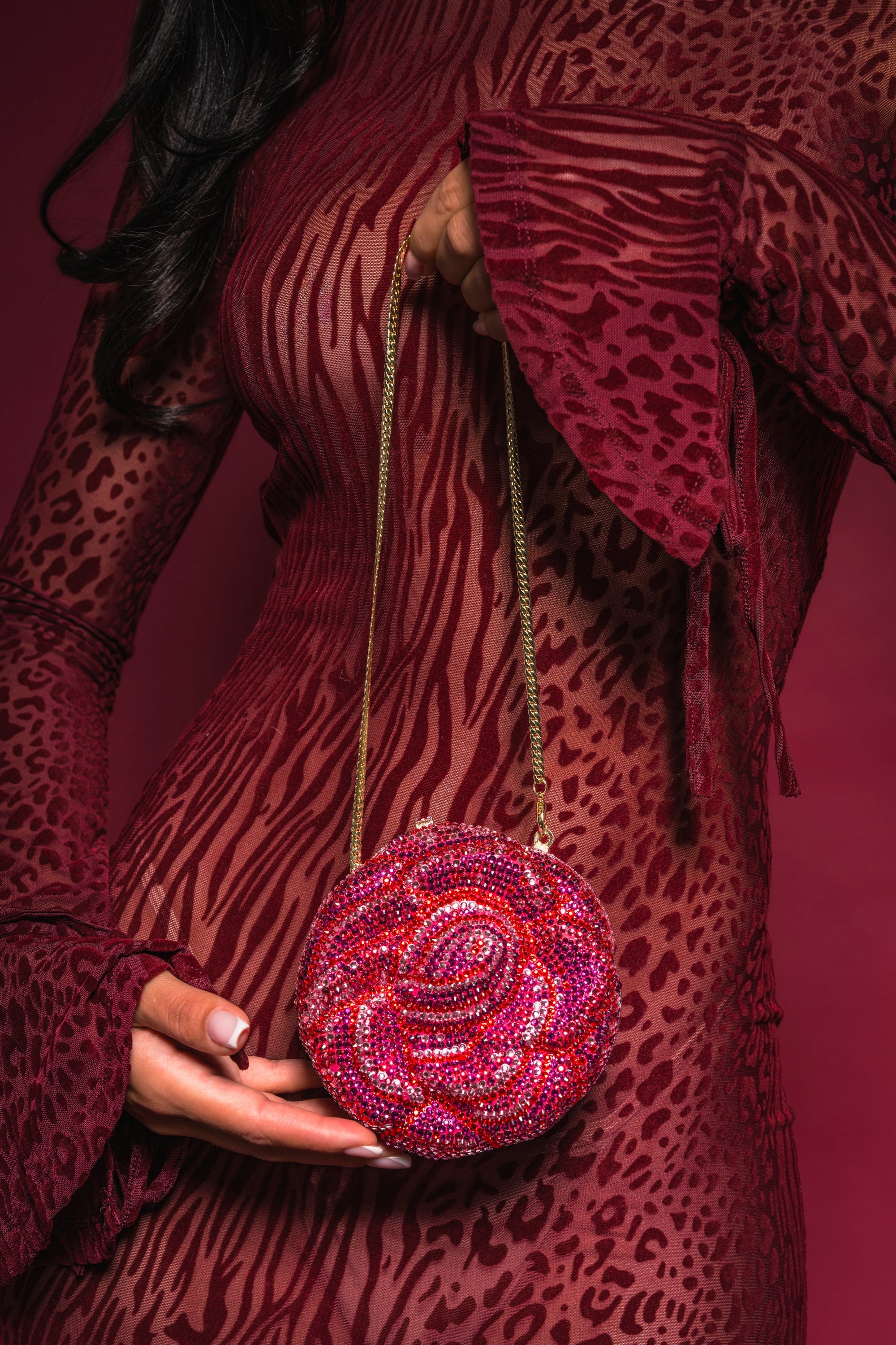 A woman holds the NOT THAT KIND OF ROSE PURSE, a sparkling rose accessory with a gold chain, set against a matching burgundy background. Only her torso and hands are visible; she's dressed in a sheer, patterned burgundy dress. 1