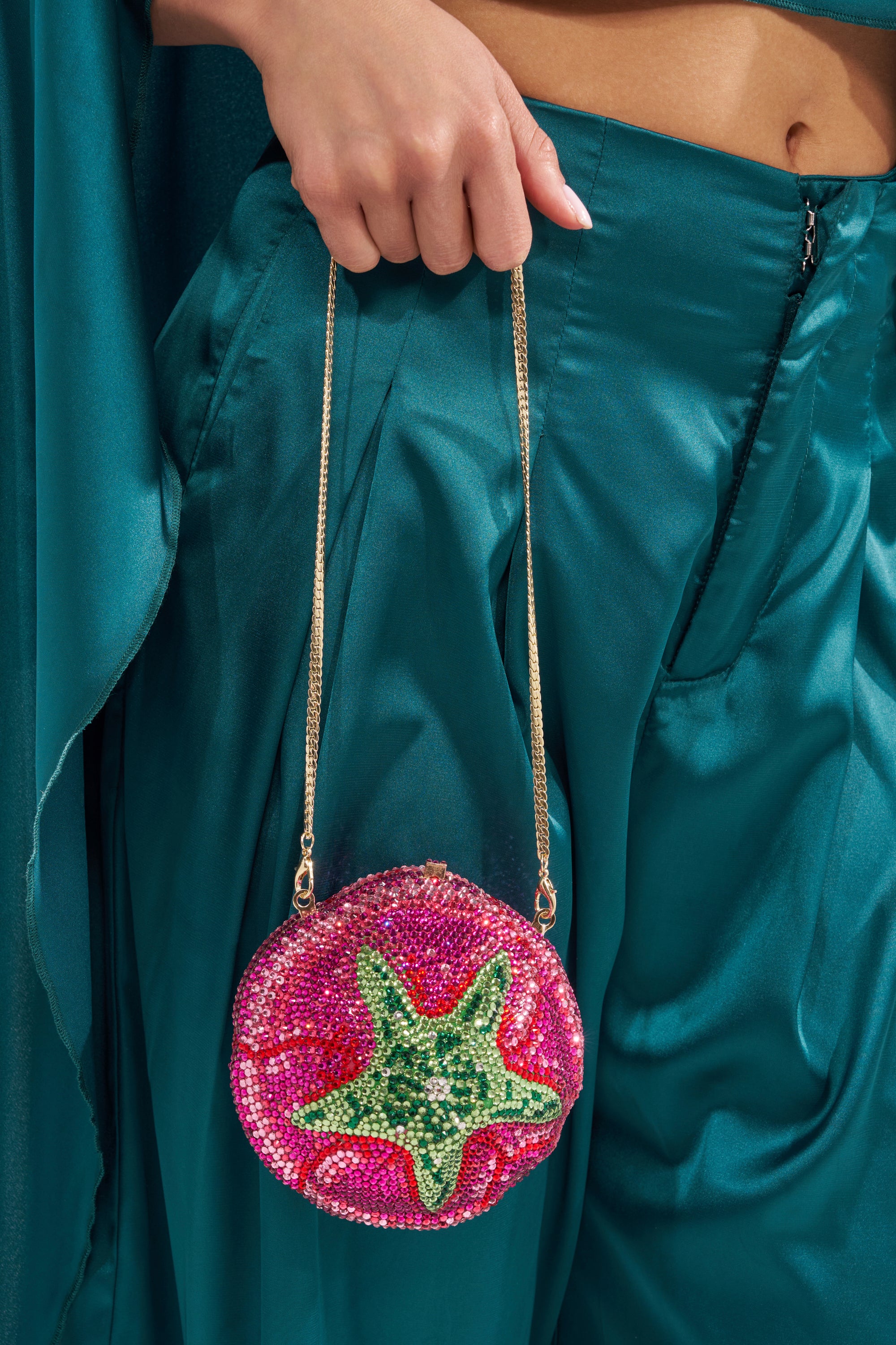 A person in teal satin pants and a matching top holds the NOT THAT KIND OF ROSE PURSE, a round rhinestone handbag with a gold chain strap, pink background, and green starfish design.