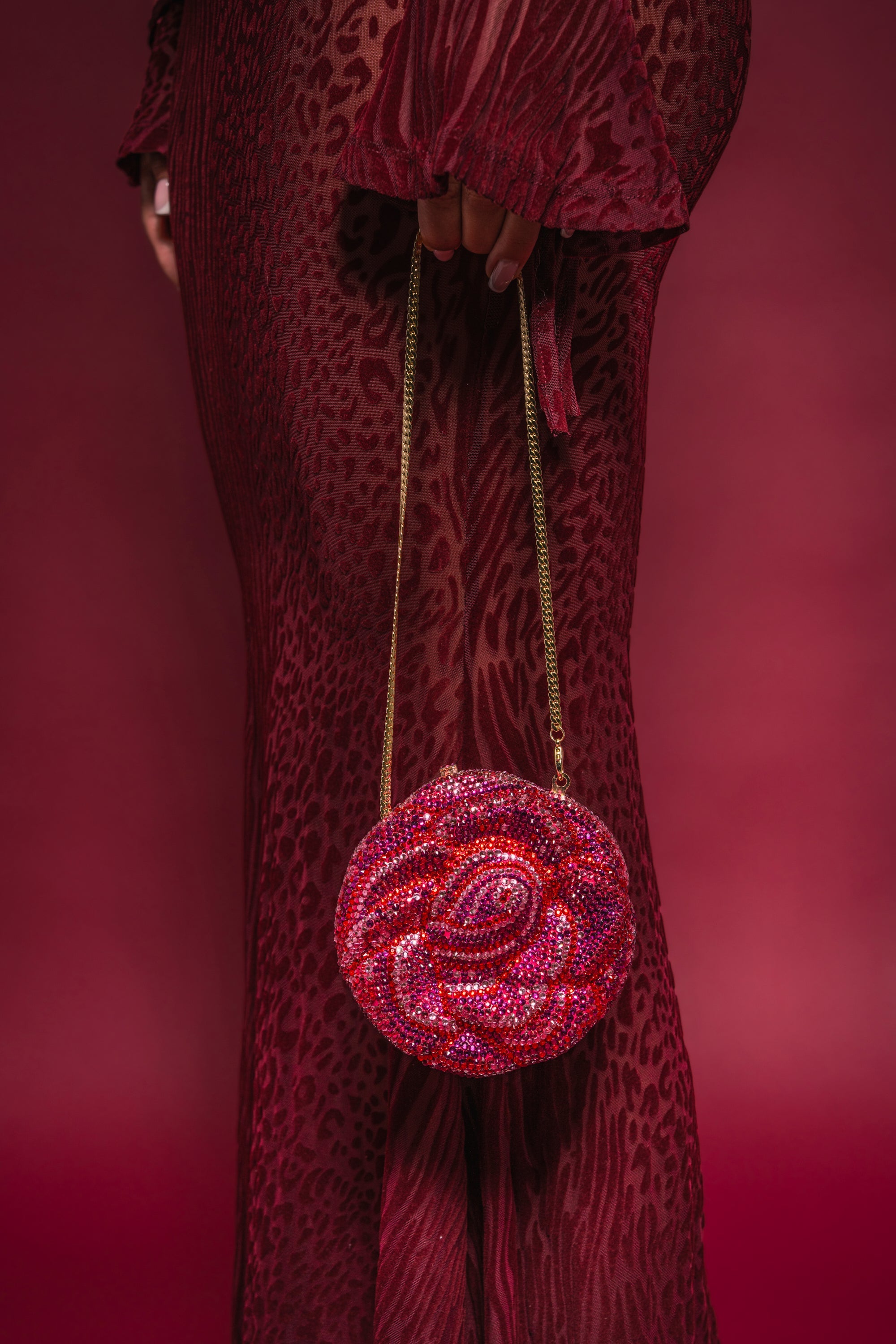 A person in a sheer red patterned dress holds the NOT THAT KIND OF ROSE PURSE, a sparkling rhinestone handbag with a gold chain handle, against a matching red background. 2